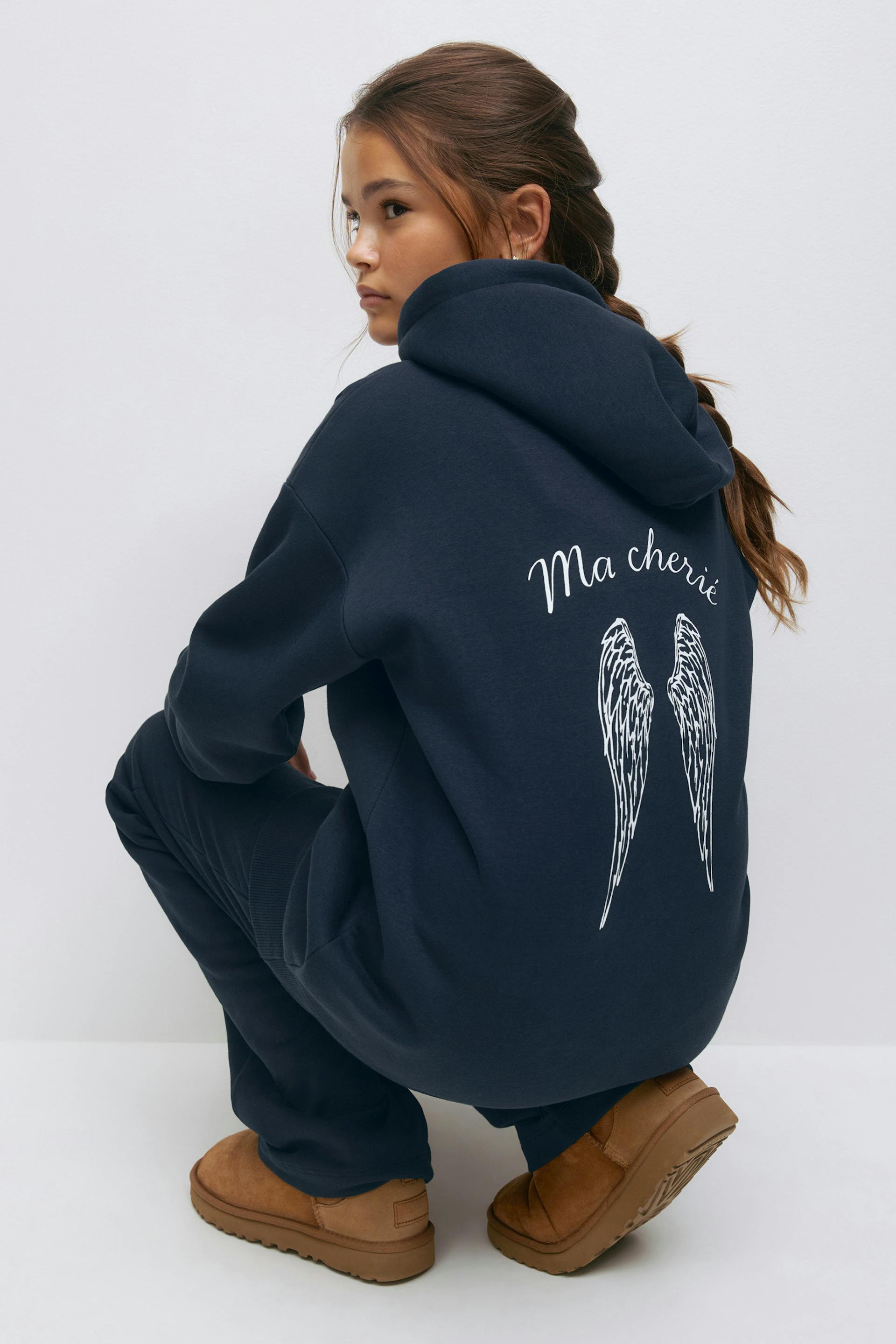 Navy oversized hooded sweatshirt featuring white "Ma chérie" script and angel wings print on the back, paired with matching navy sweatpants and tan suede slip‑on boots.