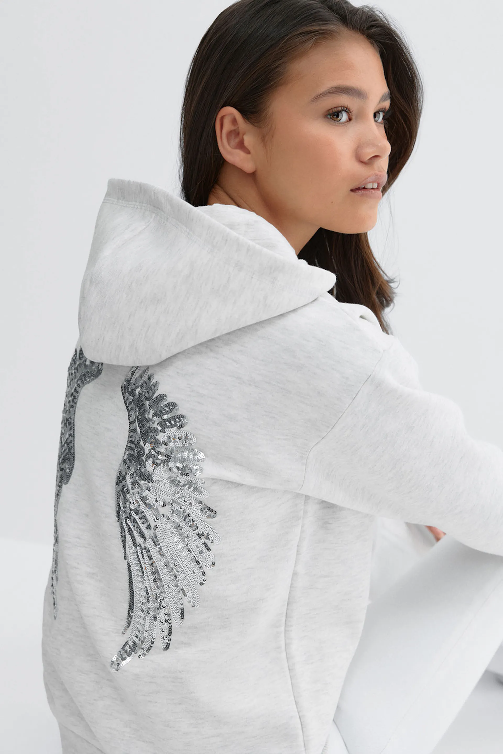 Light heather-gray hooded sweatshirt featuring a silver sequin wing motif on the back, paired with white slim-fit pants.