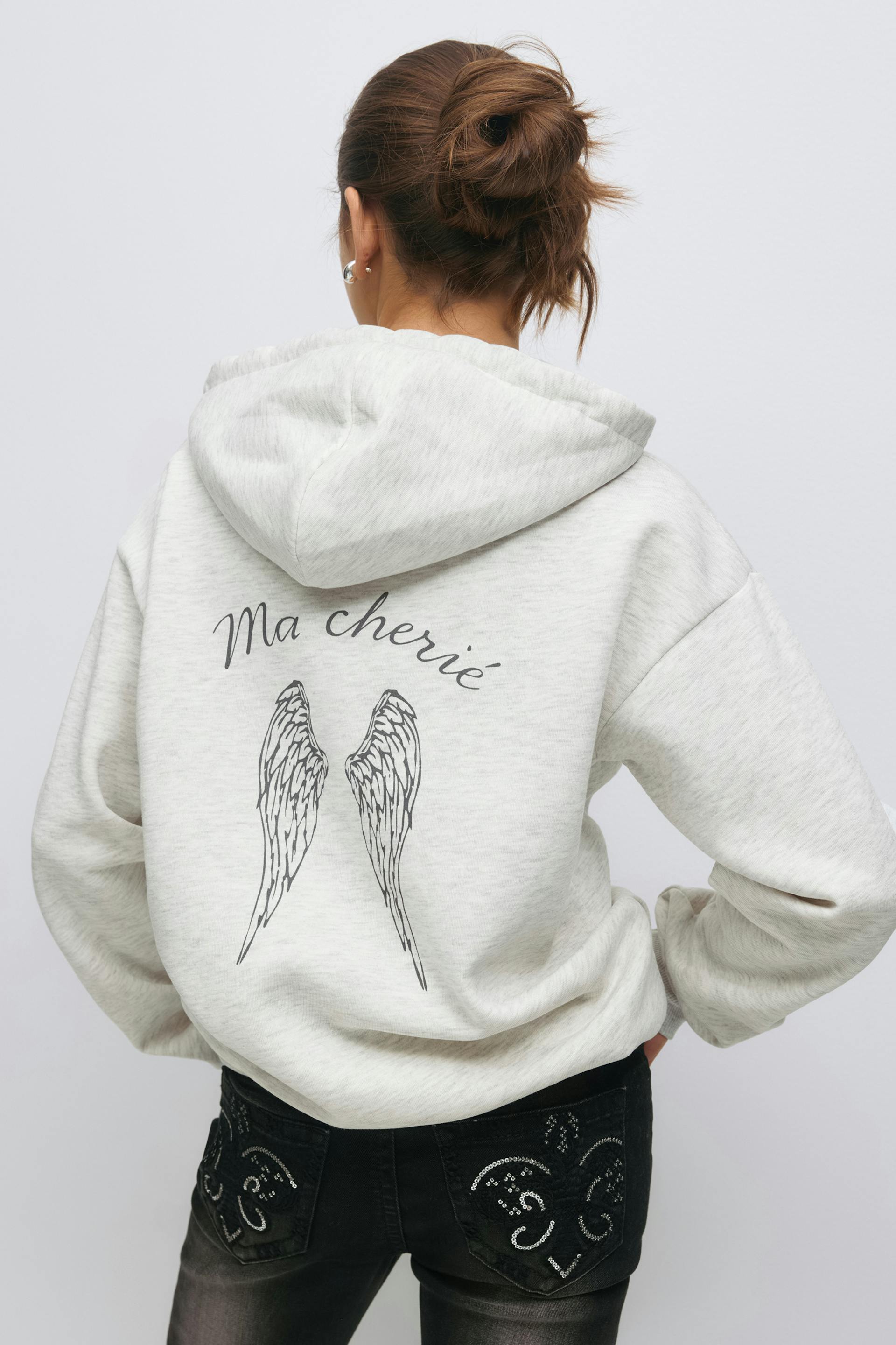Light heather-gray hooded sweatshirt with "Ma chérie" script and black angel-wing print on the back, paired with black jeans featuring decorative sequin-embellished back pockets.