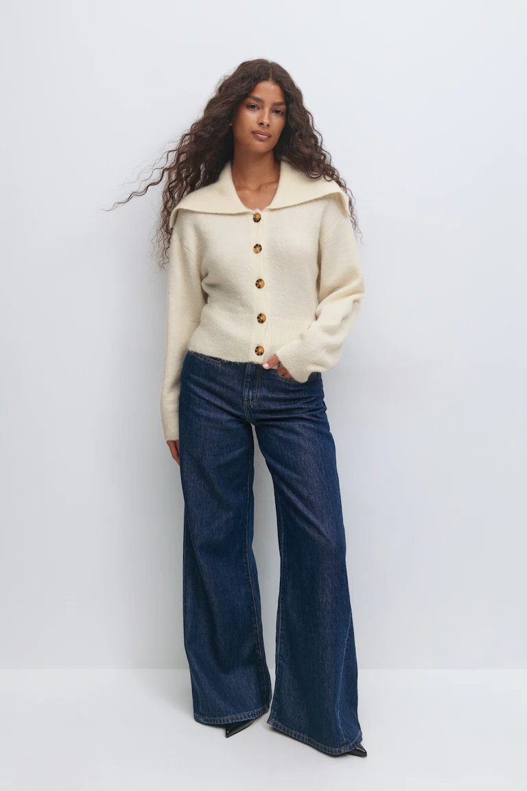 Cremefarbene Strickjacke mit breitem Kragen und schildpattfarbenen Knöpfen, kombiniert mit einer indigoblauen Jeans mit weitem Bein und schwarzen spitzen Schuhen.
