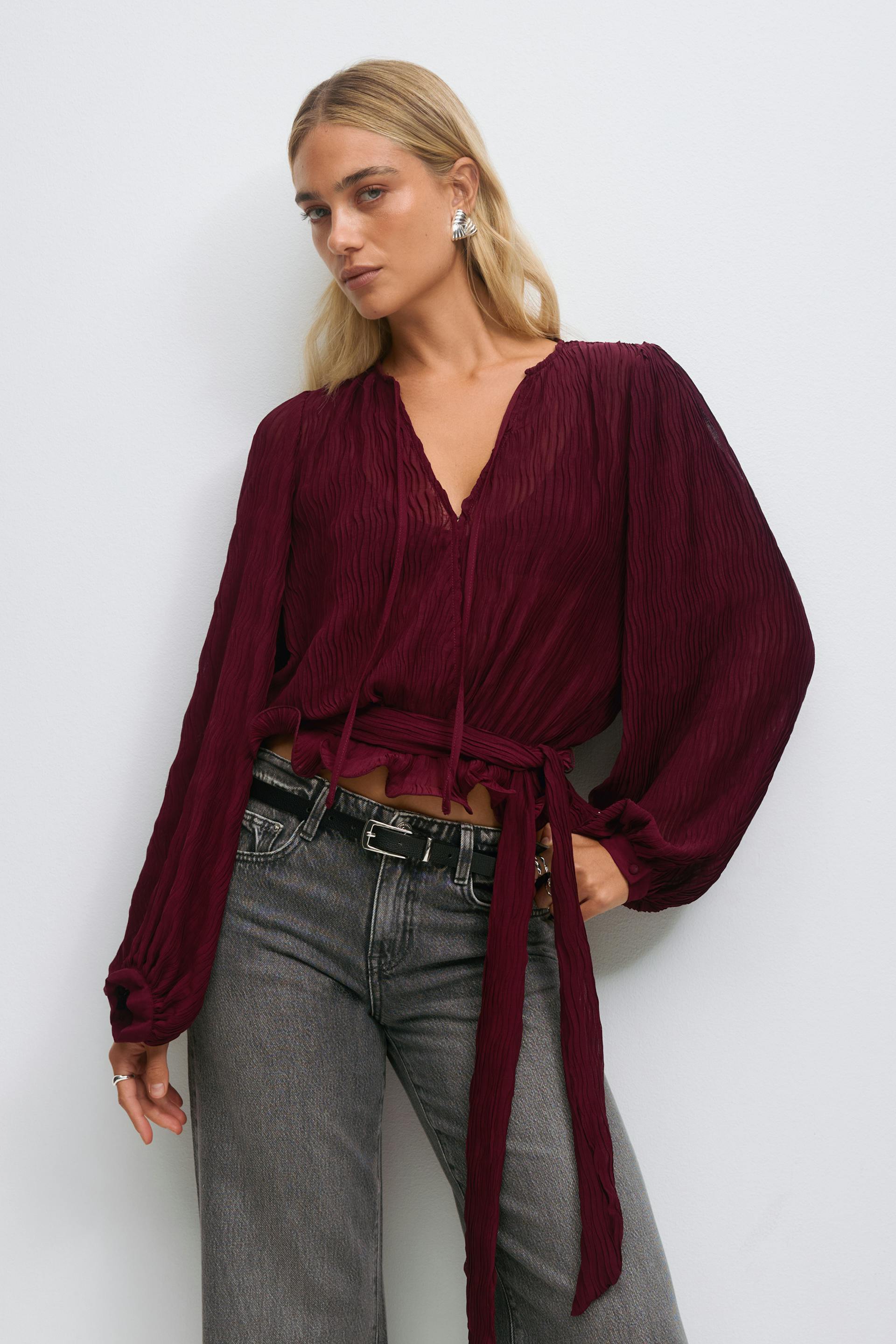 Model wearing a deep burgundy crinkled long-sleeve V-neck blouse with tie waist and a matching ruched, ruffled mini skirt, paired with black knee-high leather boots and silver hoop earrings.