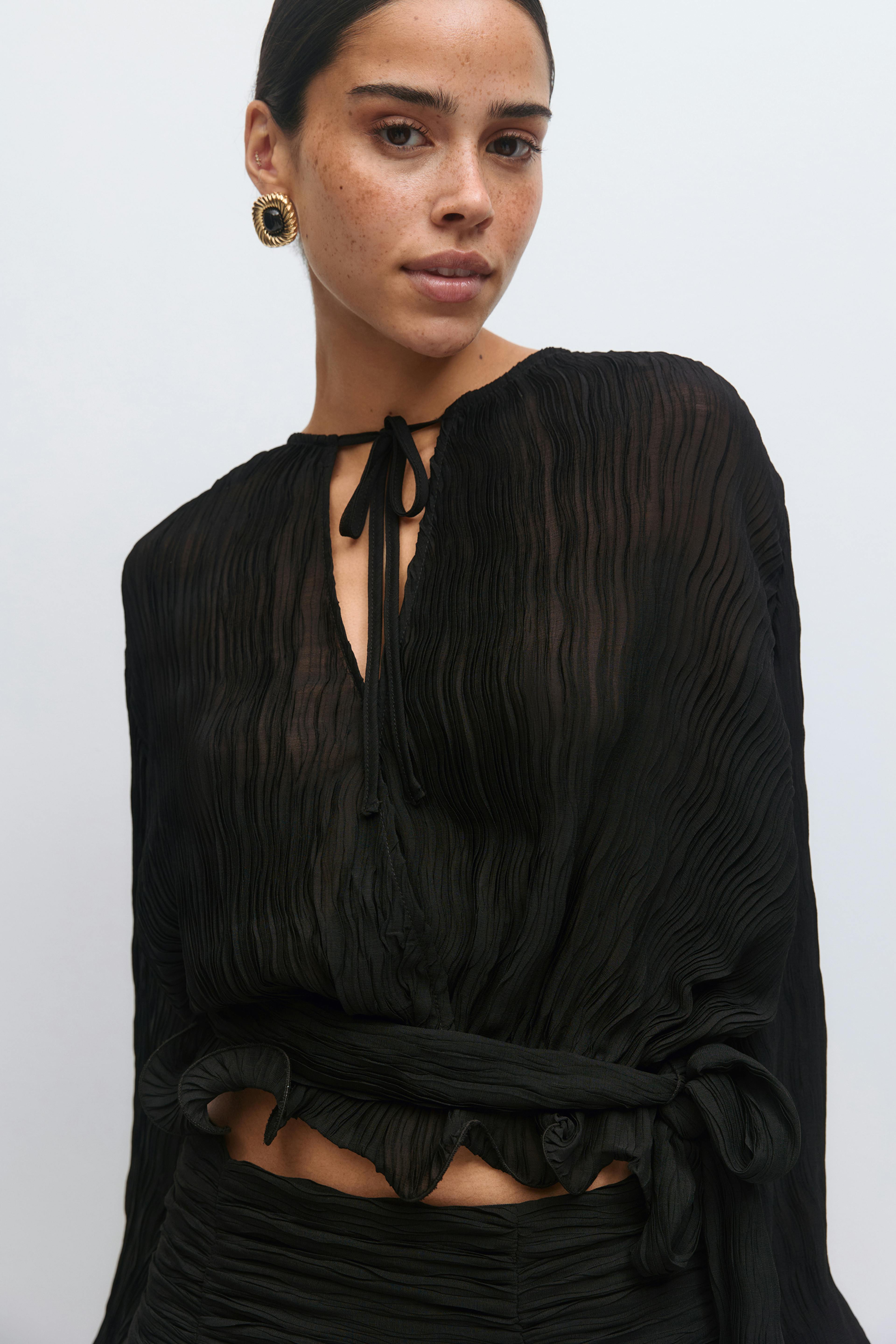 Musta laskostettu läpikuultava pitkähihainen pusero, jossa on solmio pääntieosassa, röyhelöinen röyhelöinen helma ja sivuside vyötäröllä, yhdistettynä röyhelöiseen mustaan hameeseen; kullanväriset pyöreät statement-korvakorut.