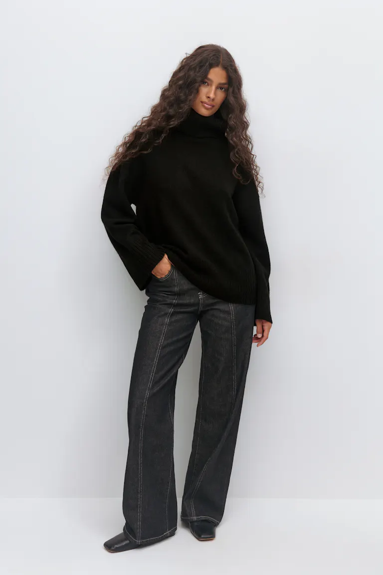 Schwarzer Oversize-Rollkragenpullover mit gerippten Bündchen und Saum, kombiniert mit dunkel gewaschenen Jeans mit weitem Bein und Naht auf der Vorderseite und schwarzen Ankle Boots mit quadratischer Fußspitze.
