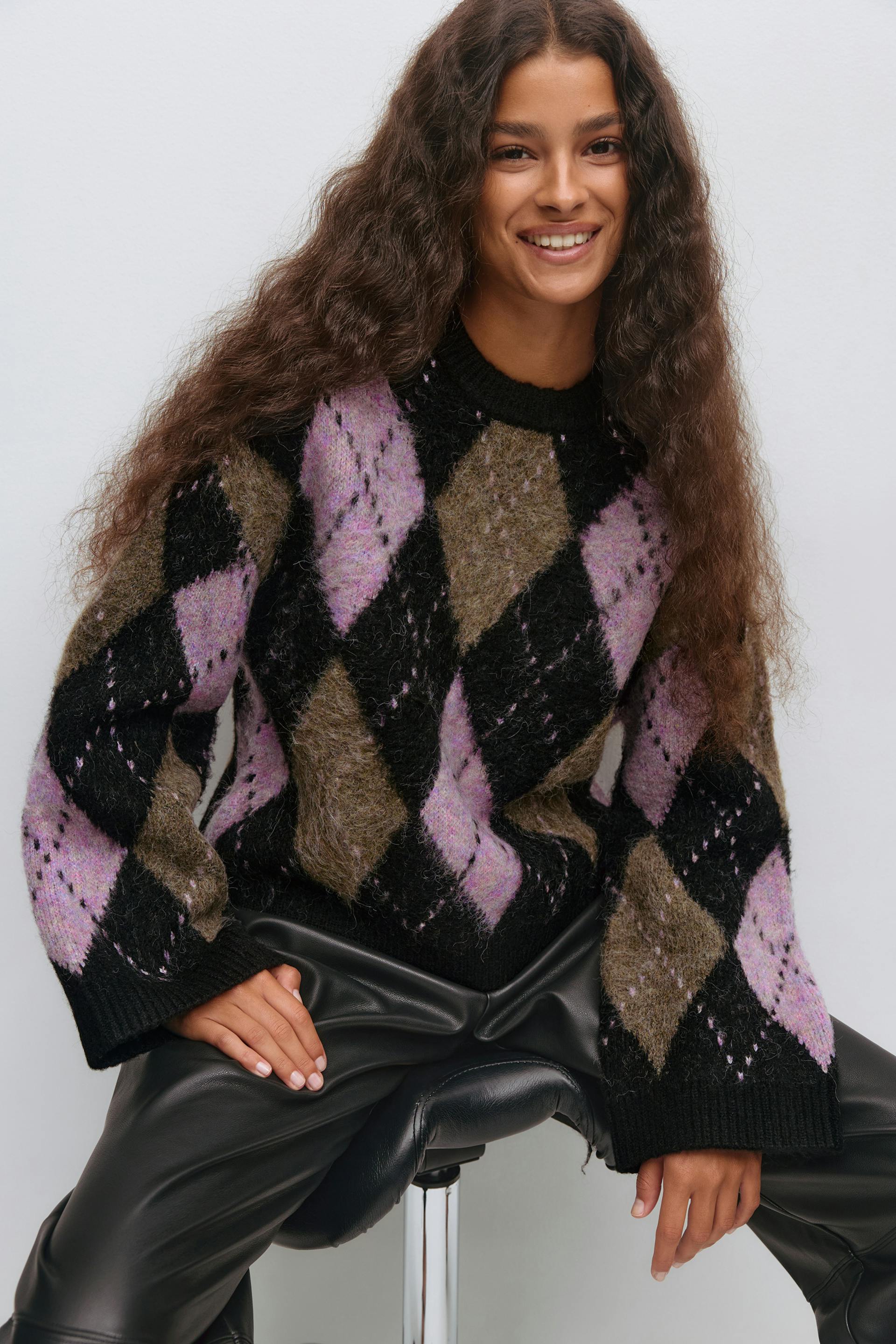 Fuzzy argyle crew-neck sweater in black, olive and pale purple with long sleeves, paired with black faux‑leather trousers.