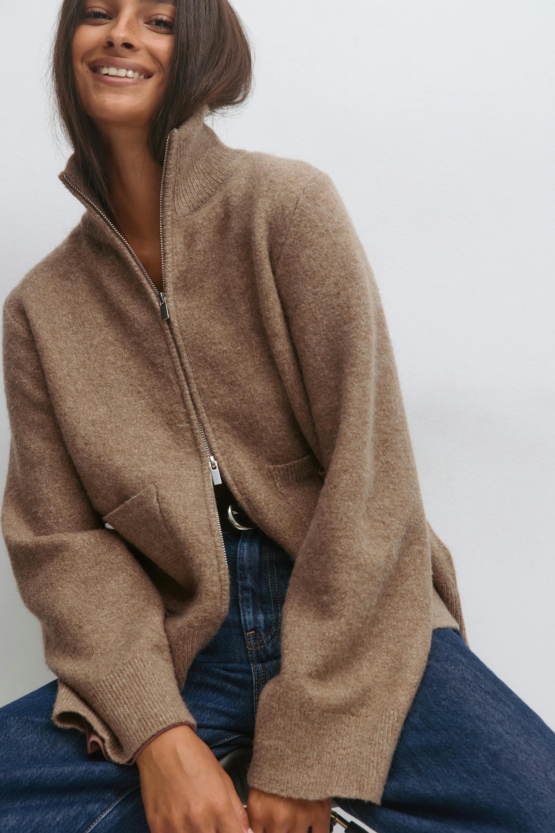 Pull en laine marron à col haut zippé et poches sur le devant, associé à un jean taille haute bleu foncé et à une ceinture noire à boucle argentée.