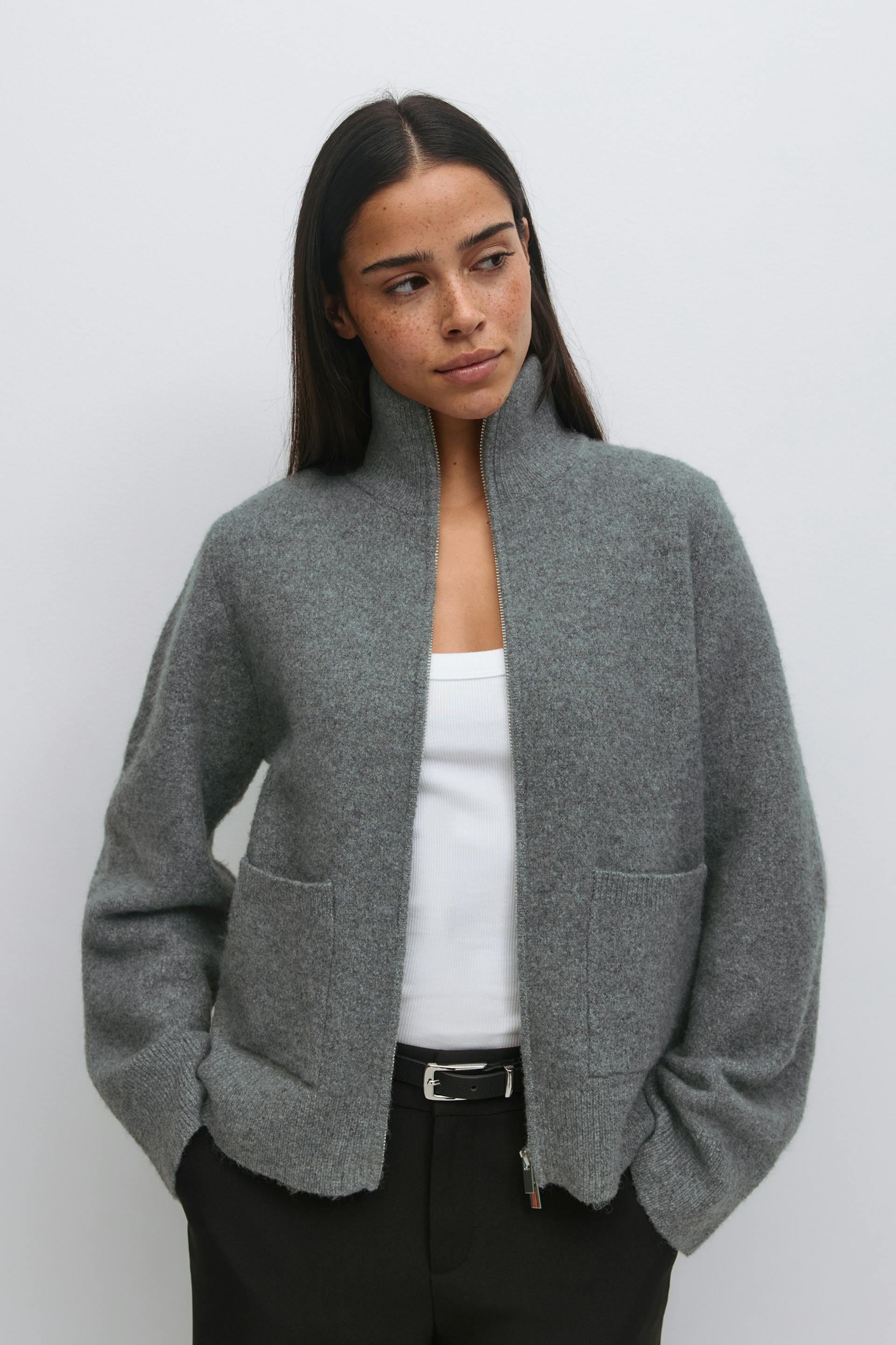 Grey zip-up wool jacket with a stand collar and front patch pockets, layered over a white ribbed tank top, paired with black trousers and a black leather belt with a silver buckle.