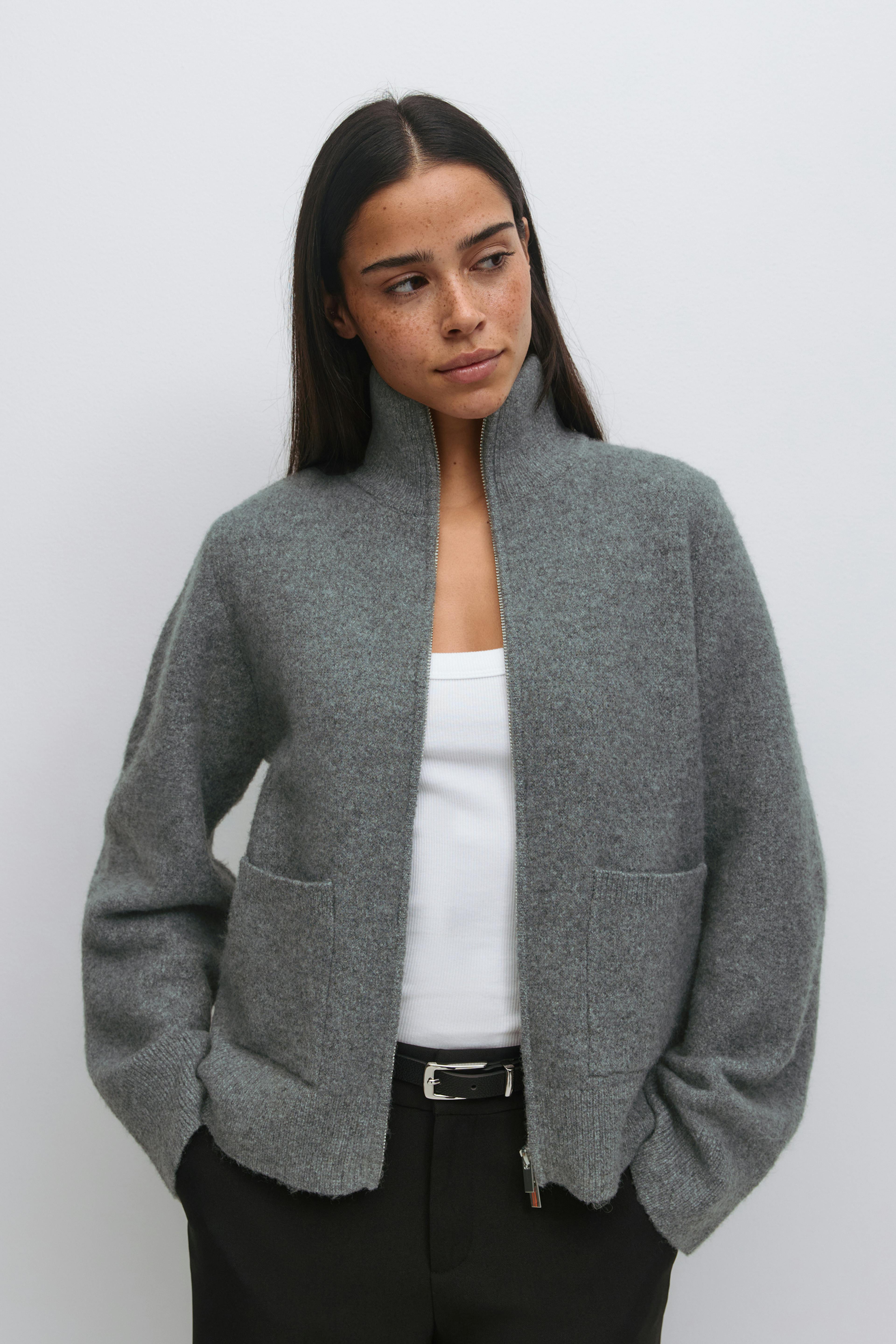 Grey zip-up wool jacket with a stand collar and front patch pockets, layered over a white ribbed tank top, paired with black trousers and a black leather belt with a silver buckle.