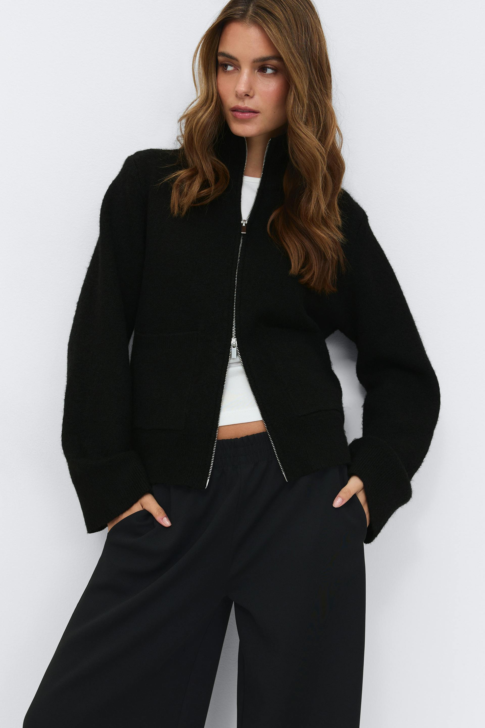 Black high‑collar zip‑up cardigan with silver zipper worn over a black ribbed crew‑neck top; small silver hoop earrings.