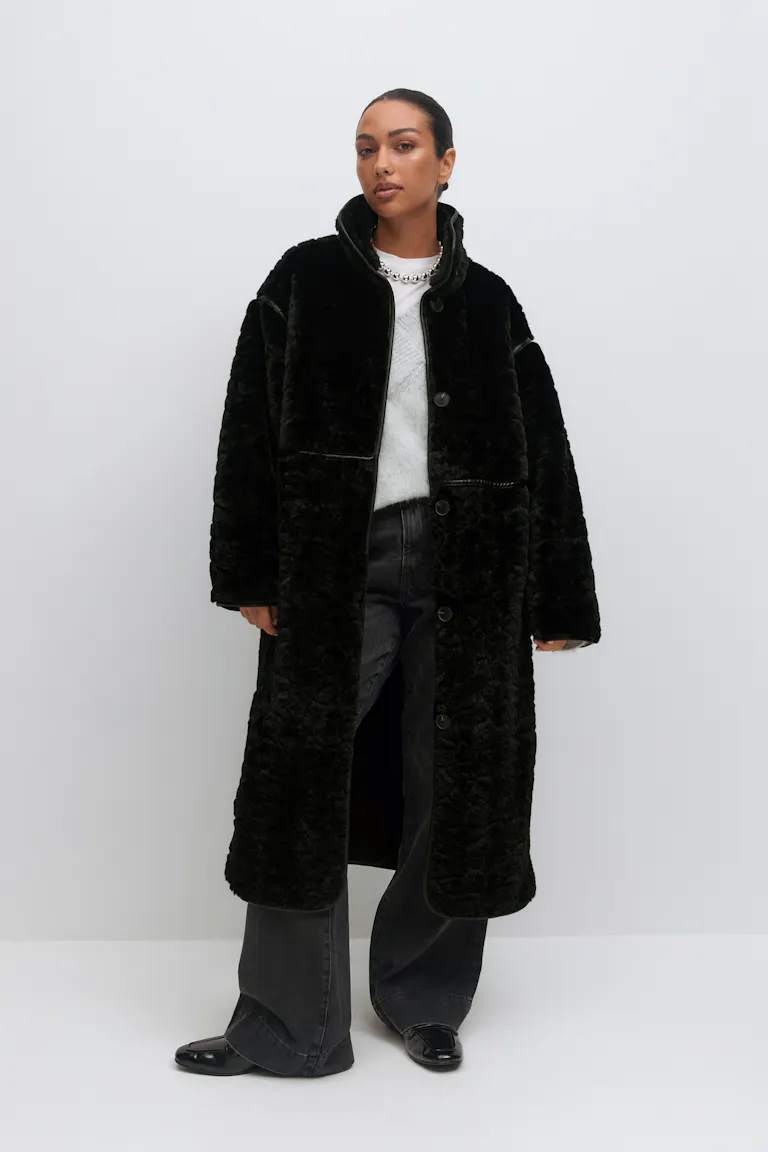 Lang sort shearling-frakke med knap foran over en hvid crewneck-sweater, sat sammen med falmede sorte jeans med brede ben, sorte lakerede loafers og en sølvkæde.