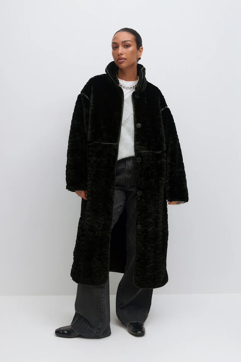 Lang sort shearling-frakke med knap foran over en hvid crewneck-sweater, sat sammen med falmede sorte jeans med brede ben, sorte lakerede loafers og en sølvkæde.