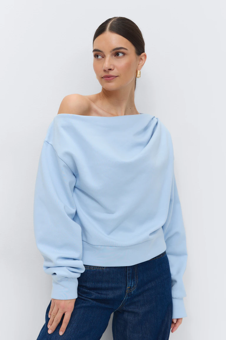 Lichtblauw gedrapeerd off-the-shoulder sweatshirt met lange mouwen gecombineerd met donkerblauwe jeans en gouden geribde hoepeloorbellen.