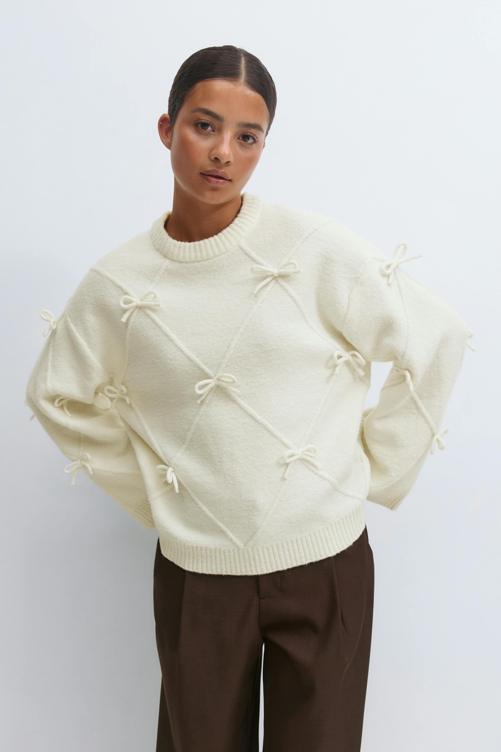 Ivory crew-neck knit sweater with ribbed neckline and hem, featuring corded diagonal diamond detailing with small bows across the body and sleeves, paired with high-waisted brown tailored trousers.