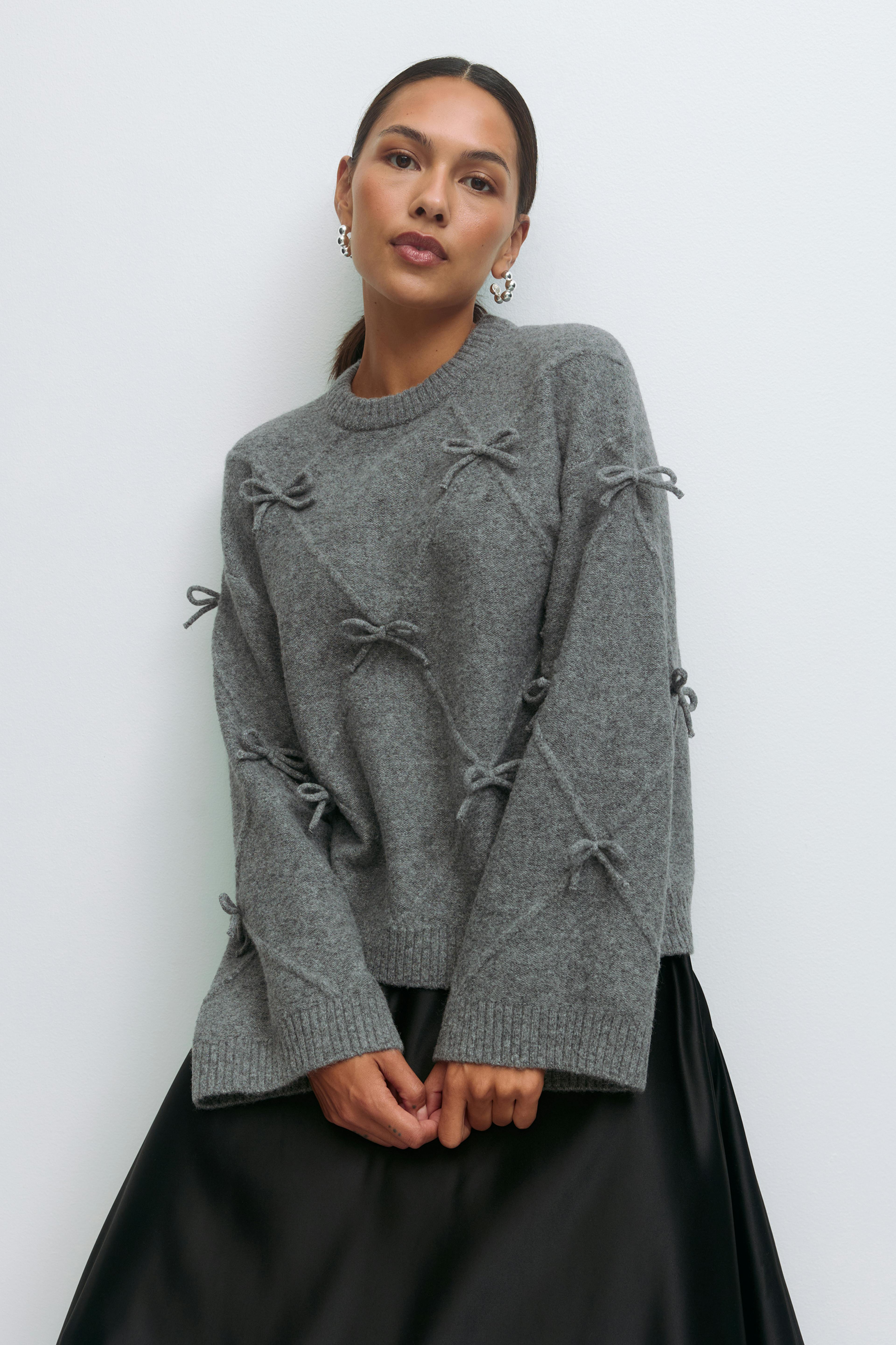 Grey crewneck wool-blend sweater with decorative tied bows and ribbed cuffs and hem, paired with a black satin skirt and silver statement hoop earrings.