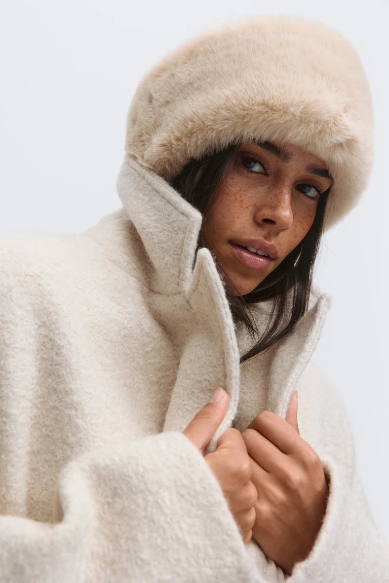 Off-white textured bouclé coat with a high stand collar, styled with a white faux‑fur hat.