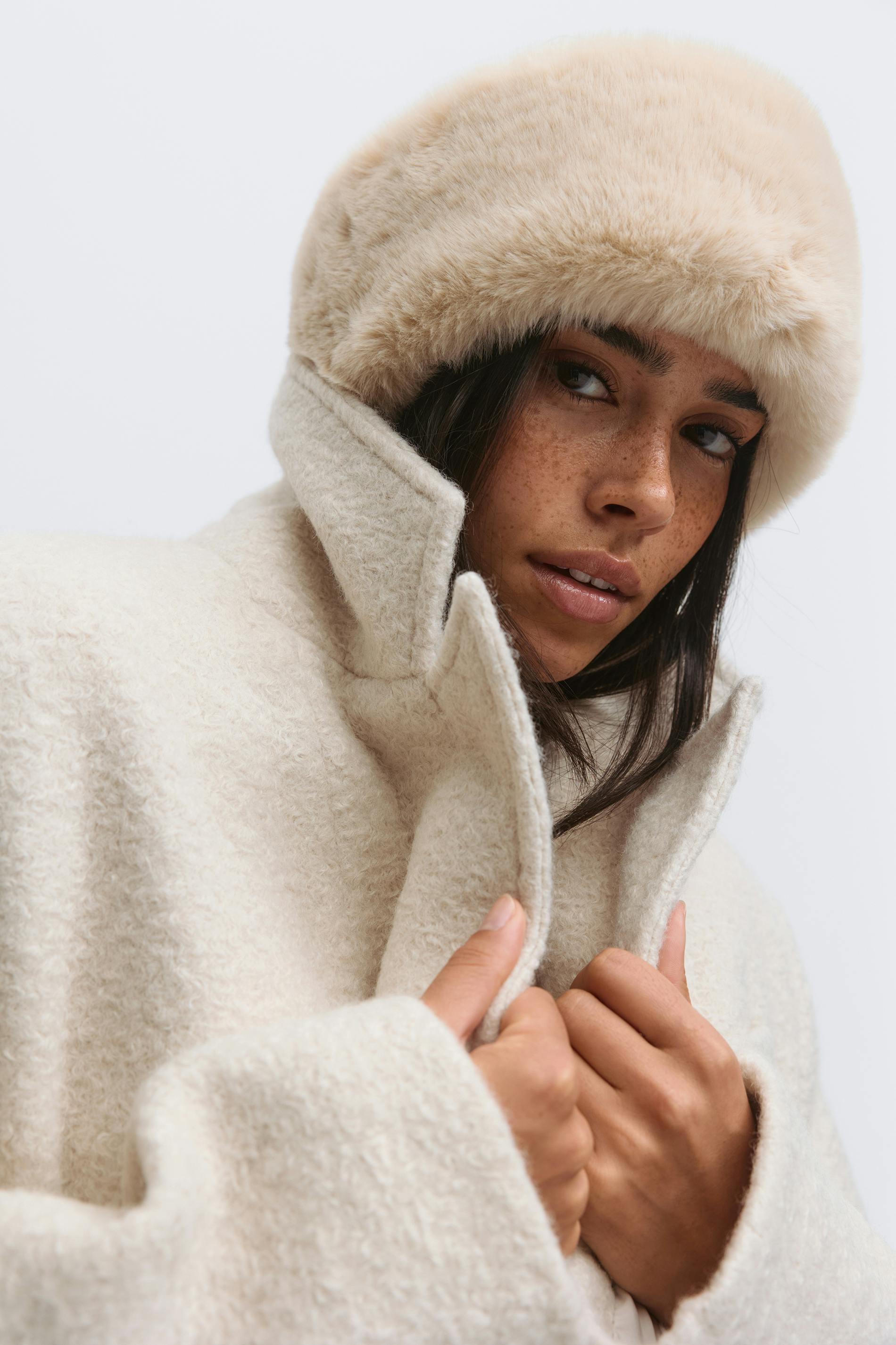 Luonnonvalkoinen kuvioitu bouclé-takki, jossa on korkea kaulus ja valkoinen tekoturkislakki.