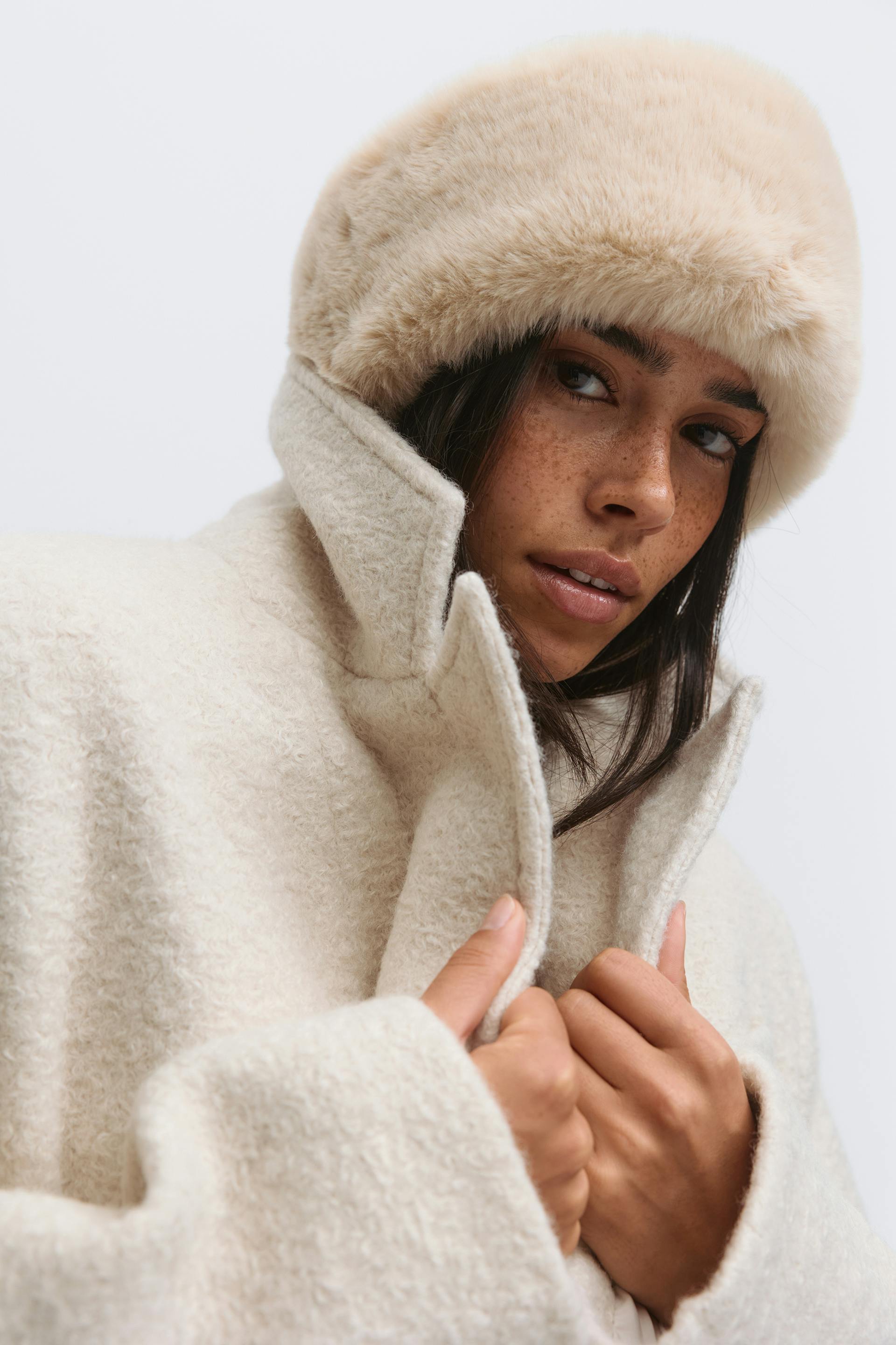 Luonnonvalkoinen kuvioitu bouclé-takki, jossa on korkea kaulus ja valkoinen tekoturkislakki.