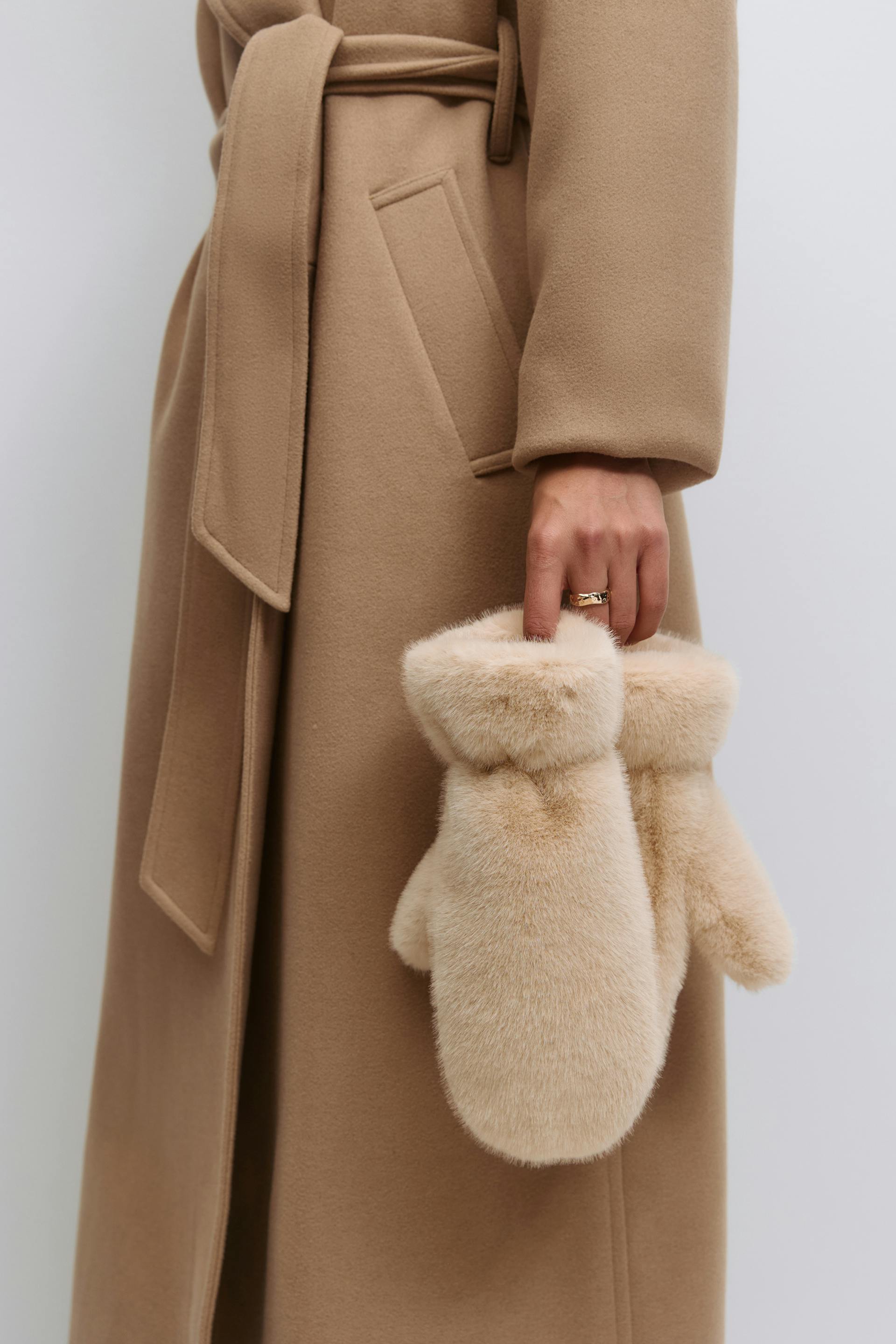 Kamelfarbener Wollmantel mit Gürtel und Leistentasche, der beigefarbene Fäustlinge aus Kunstfell trägt.