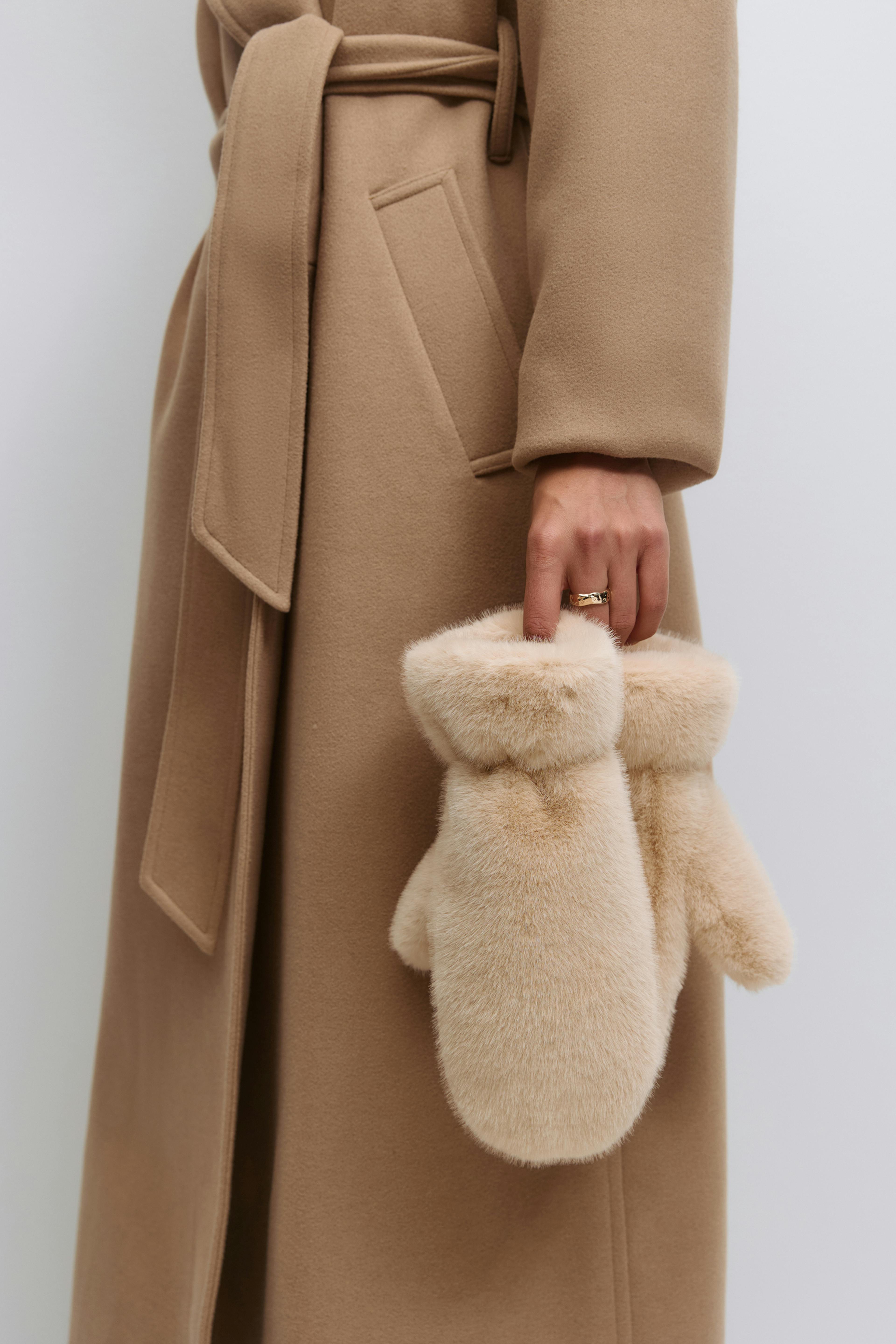 Manteau en laine ceinturé camel avec poche passepoilée, tenant des moufles en fausse fourrure beige.