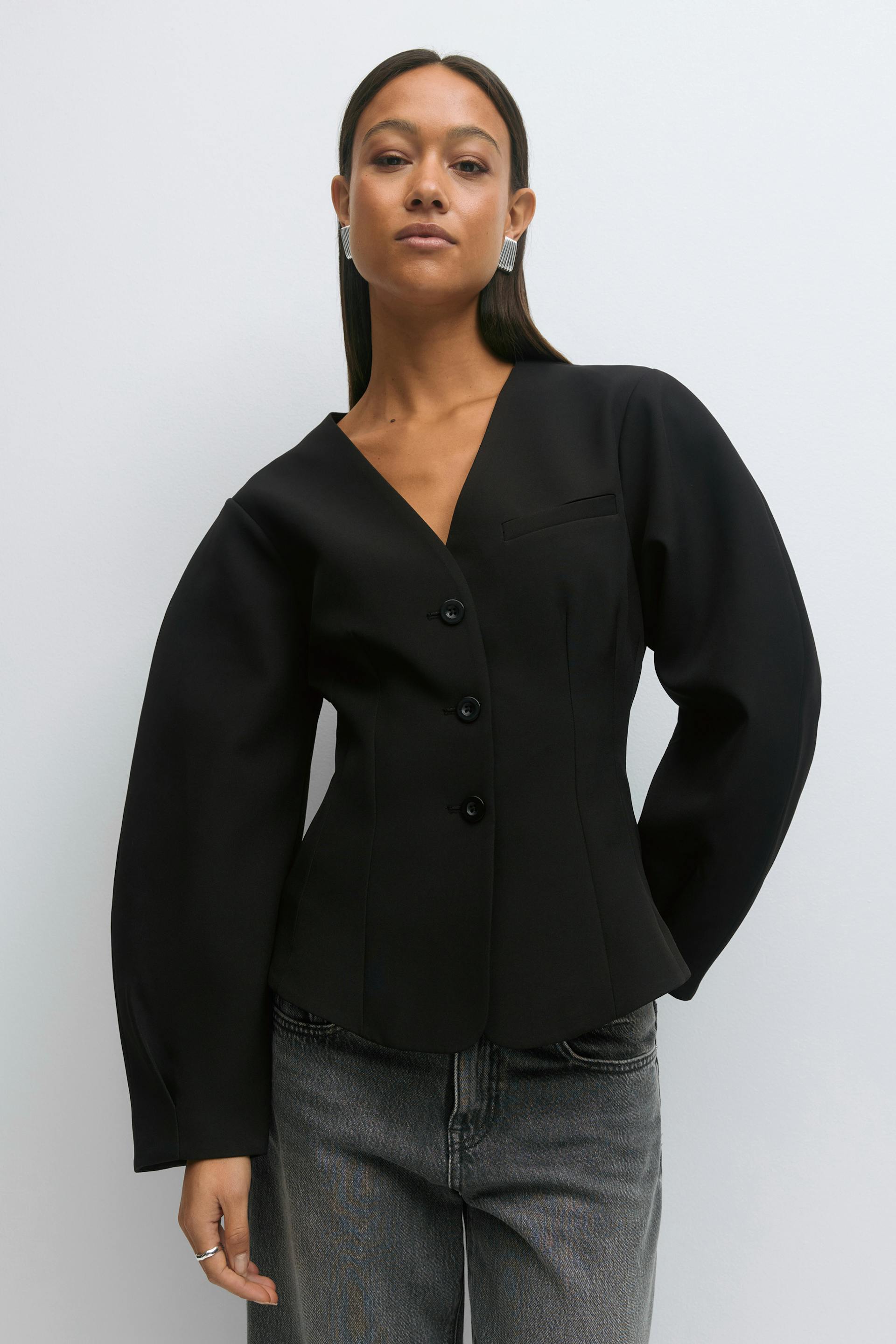 Black tailored V‑neck blazer with three-button front, fitted waist and voluminous long sleeves, paired with washed grey jeans; accessorized with silver statement earrings and a silver ring.