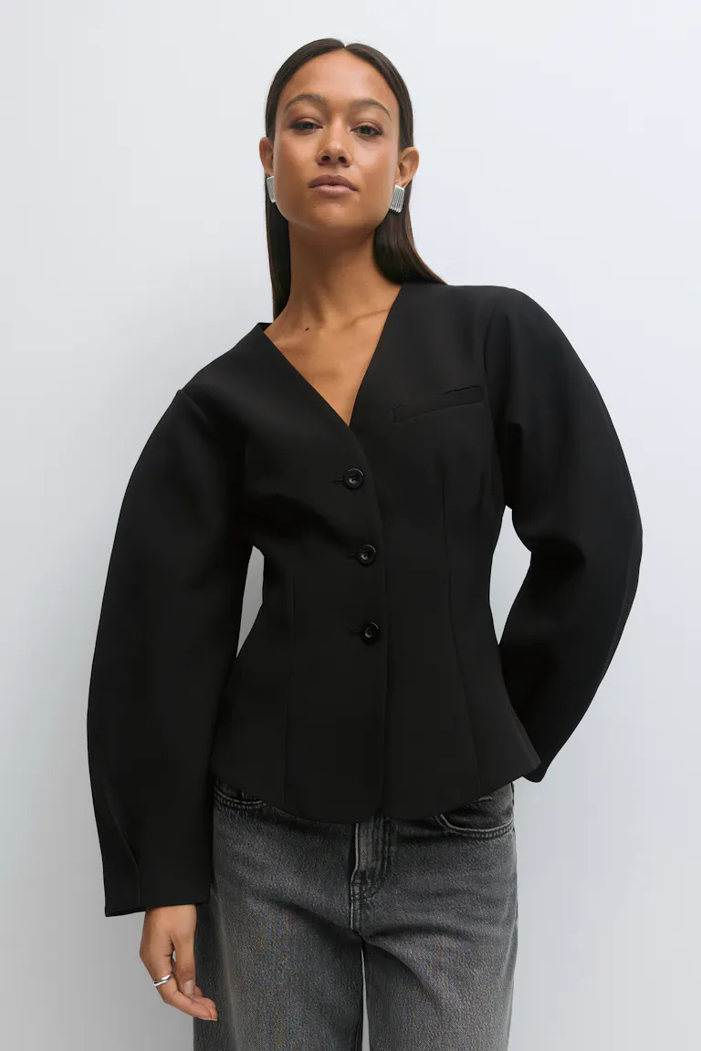 Schwarzer, taillierter Blazer mit V-Ausschnitt, drei Knöpfen, taillierter Passform und voluminösen langen Ärmeln, kombiniert mit einer verwaschenen grauen Jeans; dazu silberne Statement-Ohrringe und ein silberner Ring.