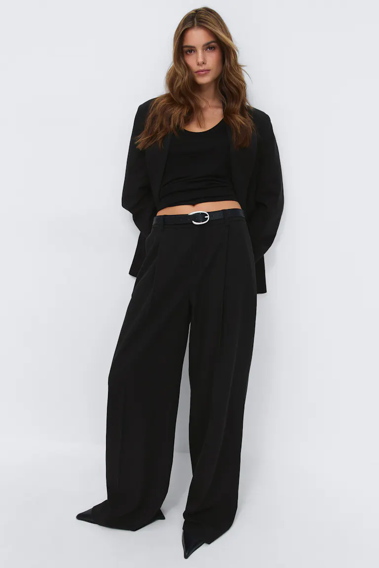 Kvinde iført en sort oversize blazer over en sort kort tanktop, med højtaljede sorte bukser med brede ben, et sort bælte med et ovalt sølvspænde og spidse sorte hæle.
