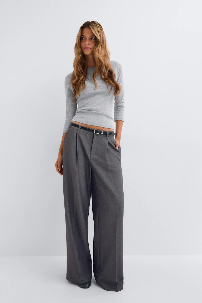 Houtskoolgrijze oversized blazer met bijpassende wijde broek met hoge taille, lichtgrijze cropped tank, zwarte leren riem, zwarte schoenen met spitse tenen en gelaagde hangerkettingen.