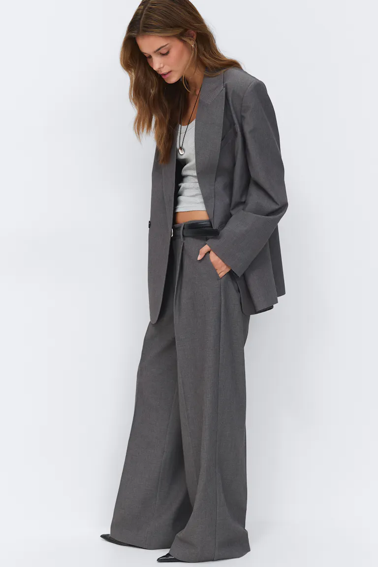 Houtskoolgrijze oversized blazer met bijpassende wijde broek met hoge taille, lichtgrijze cropped tank, zwarte leren riem, zwarte schoenen met spitse tenen en gelaagde hangerkettingen.
