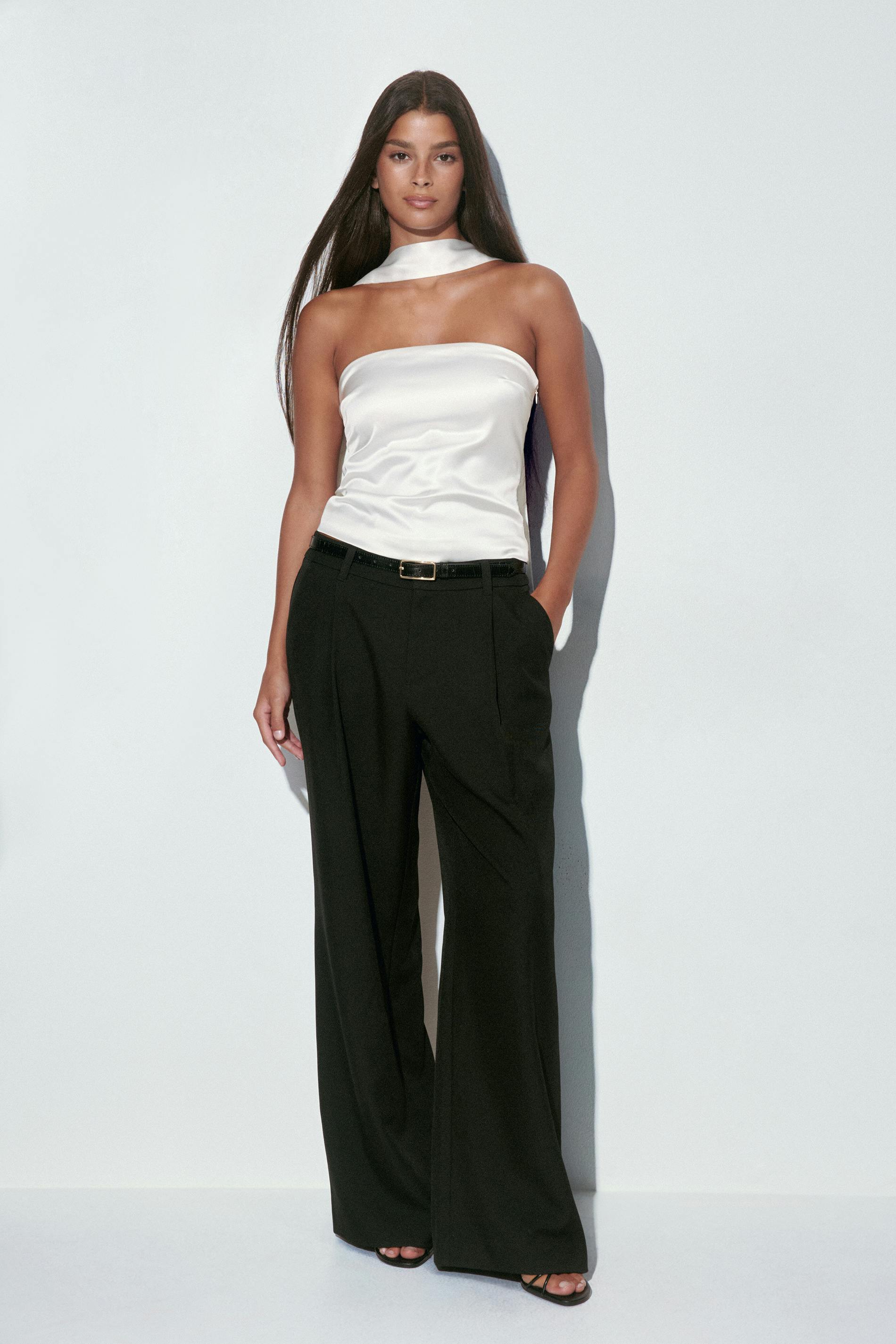 White satin strapless bandeau top with attached narrow neck scarf, paired with high-waisted black tailored wide-leg trousers, a slim black belt with gold buckle, and black strappy open-toe sandals.