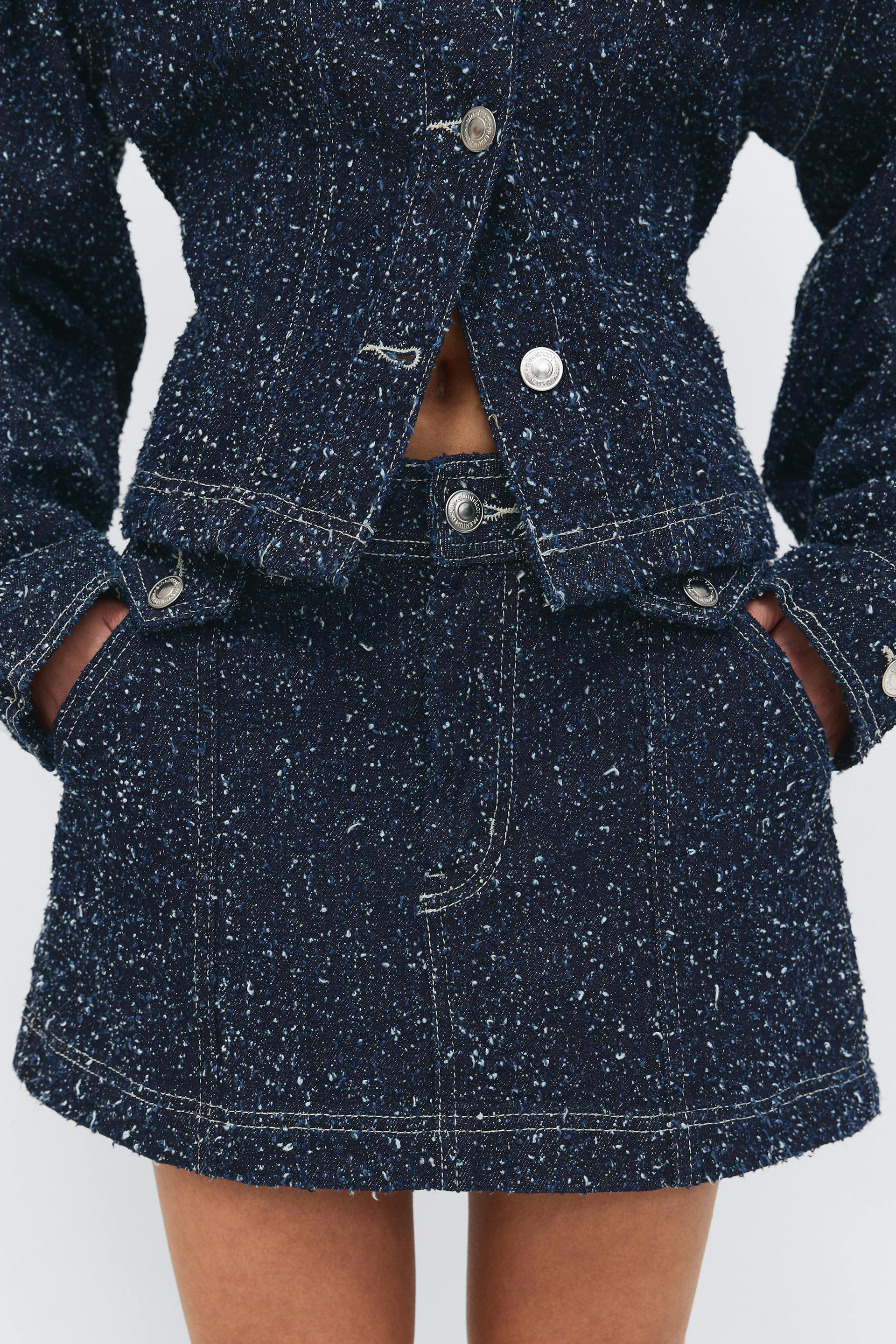 Cropped navy speckled tweed jacket with silver buttons and contrast stitching, paired with a matching high‑waisted mini skirt featuring front pockets and button details.