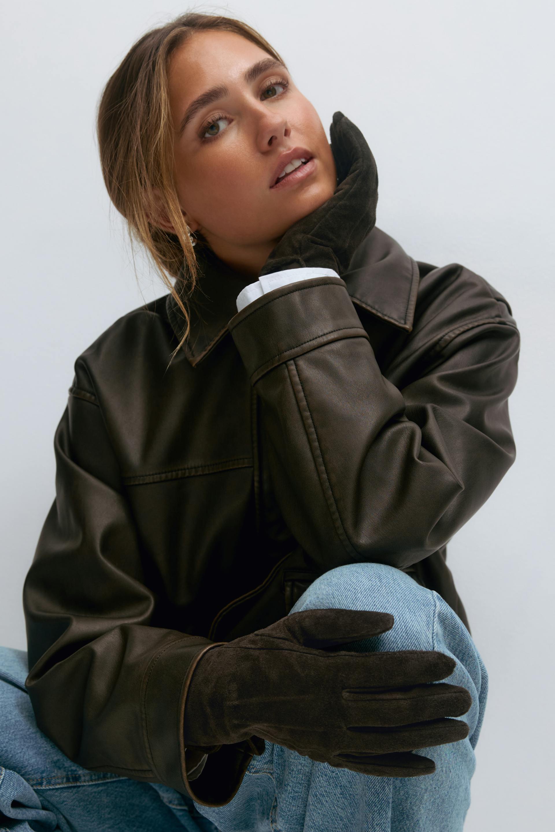 Dark brown leather collared jacket with a white‑cuffed shirt, paired with light blue jeans and dark brown suede gloves.