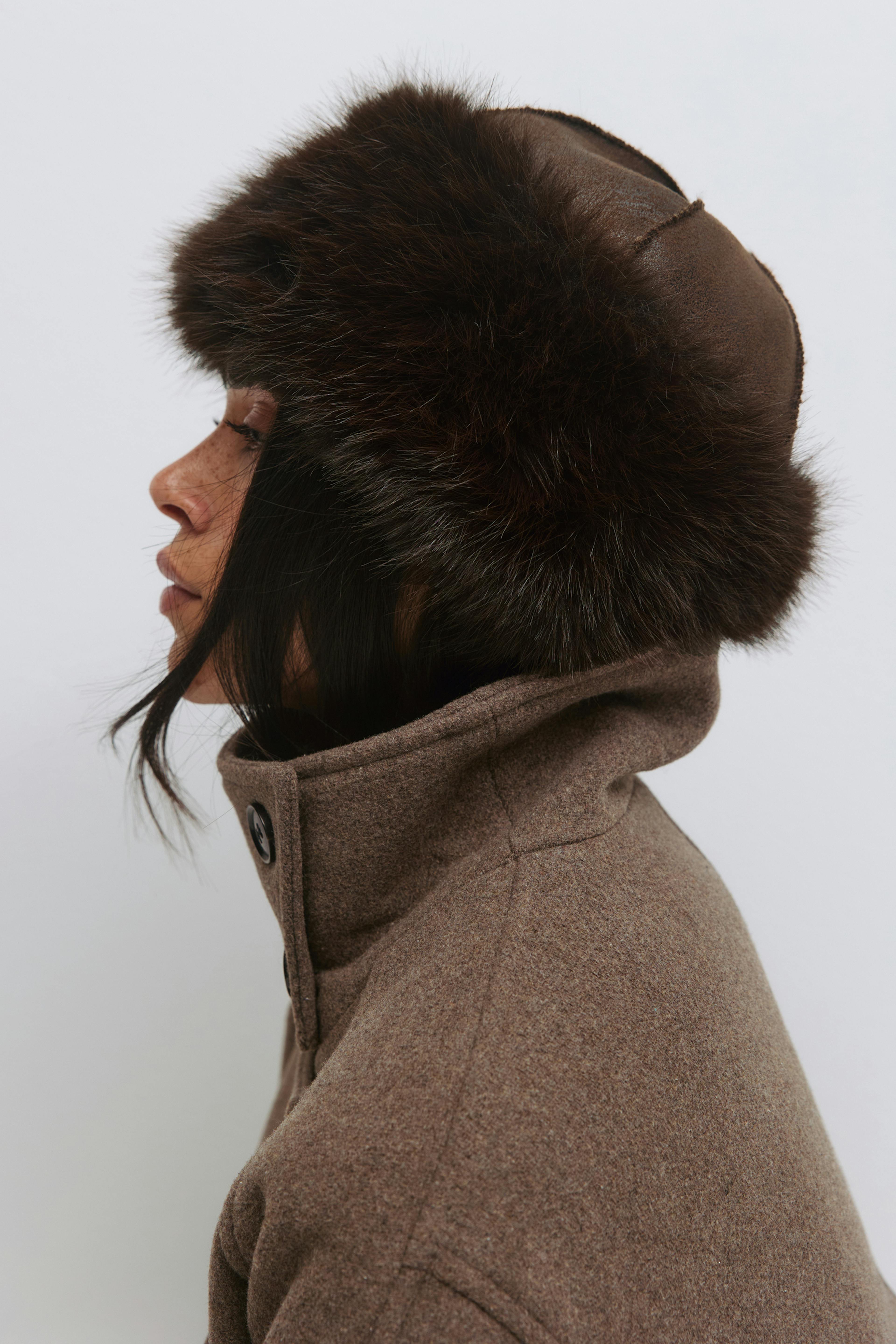 Tummanruskea turkisverhoiltu trapperihattu yhdistettynä ruskeaan korkeakauluksiseen villatakkiin, jossa on napitettu kaulalista.