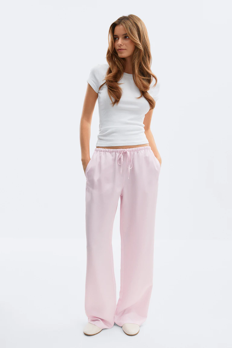 Wit T-shirt met korte mouwen en ronde hals in combinatie met een lichtroze broek met wijde pijpen en ivoorkleurige slippertjes.
