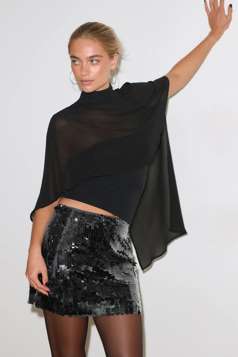Durchsichtiges schwarzes, asymmetrisches Cape-Top mit Rundhalsausschnitt, das über ein schwarzes, tailliertes Crop-Top gestülpt wird, kombiniert mit einem schwarzen, mit Pailletten besetzten Minirock, einer schwarzen, durchsichtigen Strumpfhose und großen, silbernen Ohrringen.