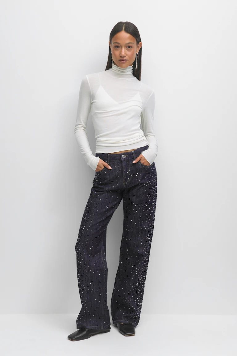 Das Modell trägt einen weißen, langärmeligen Rollkragenpullover über einem weißen Camisole, eine dunkle, indigoblaue Jeans mit weitem Bein und vereinzelten silberfarbenen Verzierungen, schwarze, quadratische Halbschuhe und lange, tropfenförmige Ohrringe aus Strass.
