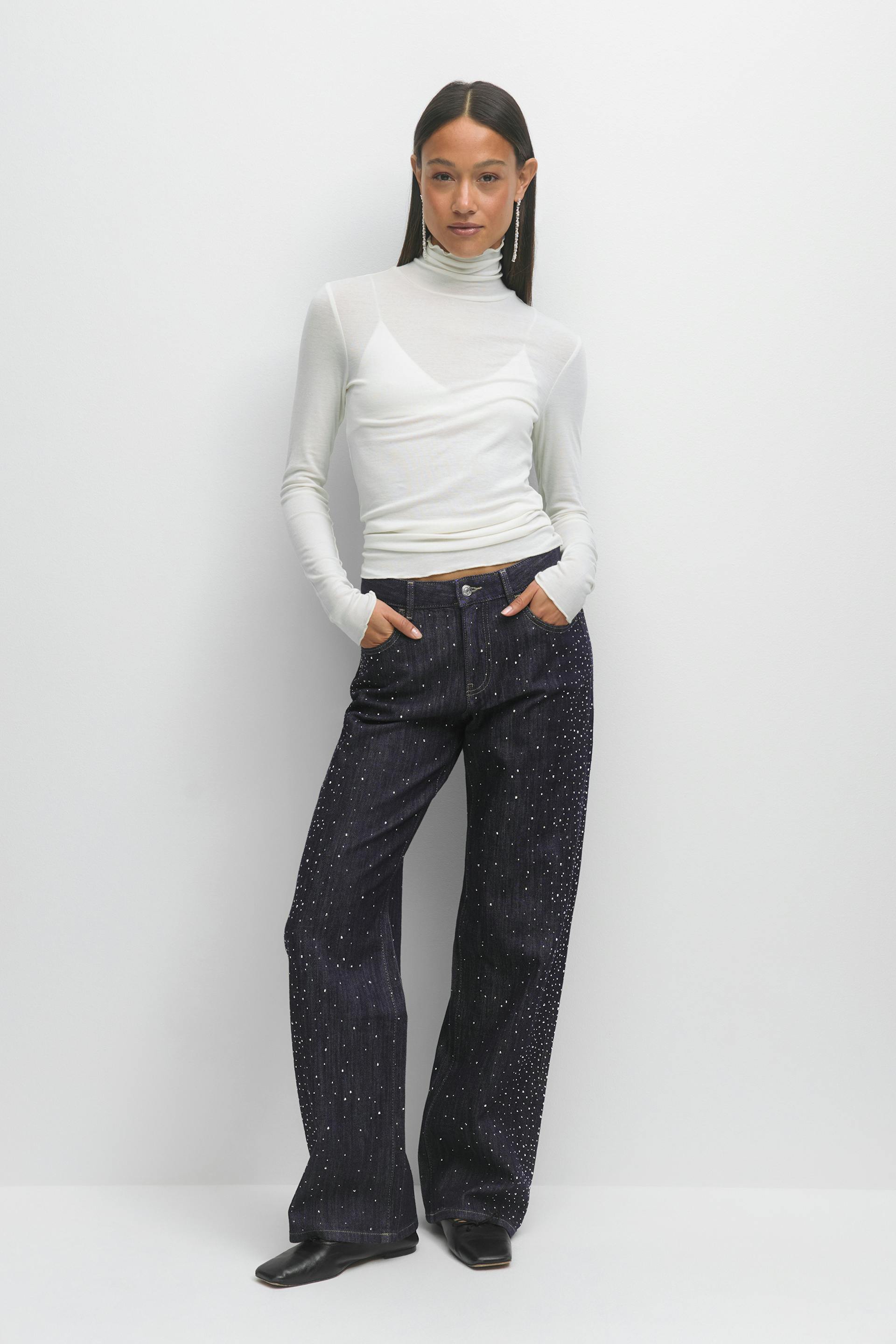 Das Modell trägt einen weißen, langärmeligen Rollkragenpullover über einem weißen Camisole, eine dunkle, indigoblaue Jeans mit weitem Bein und vereinzelten silberfarbenen Verzierungen, schwarze, quadratische Halbschuhe und lange, tropfenförmige Ohrringe aus Strass.