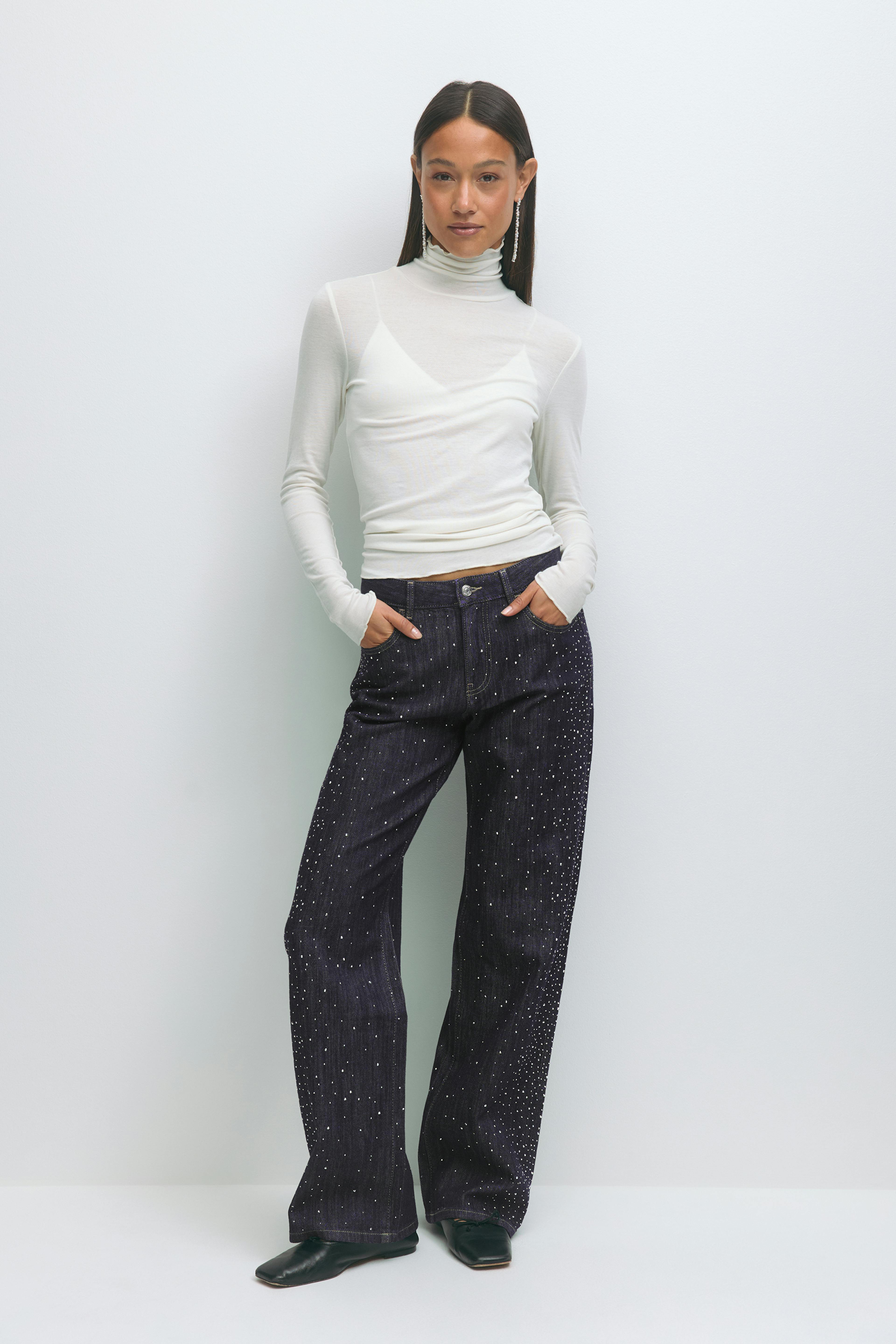 Weißer, langärmeliger Rollkragenpullover über einem weißen Mieder; dunkle Jeans mit weitem Bein und hoher Taille und verstreuten Metallnieten; schwarze, spitze Schuhe; lange Ohrringe.