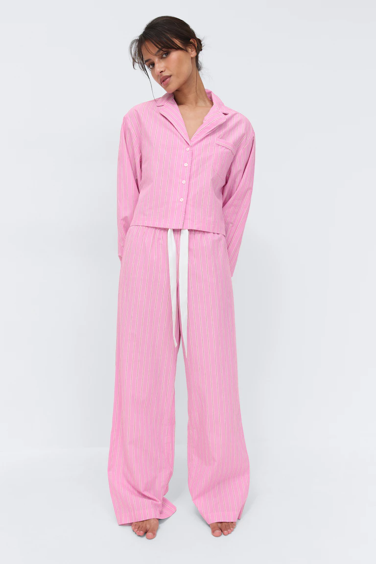 Zweiteiliges Pyjama-Set in Rosa mit vertikalen Nadelstreifen: Langärmeliges Hemd mit Kragen und Brusttasche, gepaart mit einer Hose mit weitem Bein und weißer Schleife.