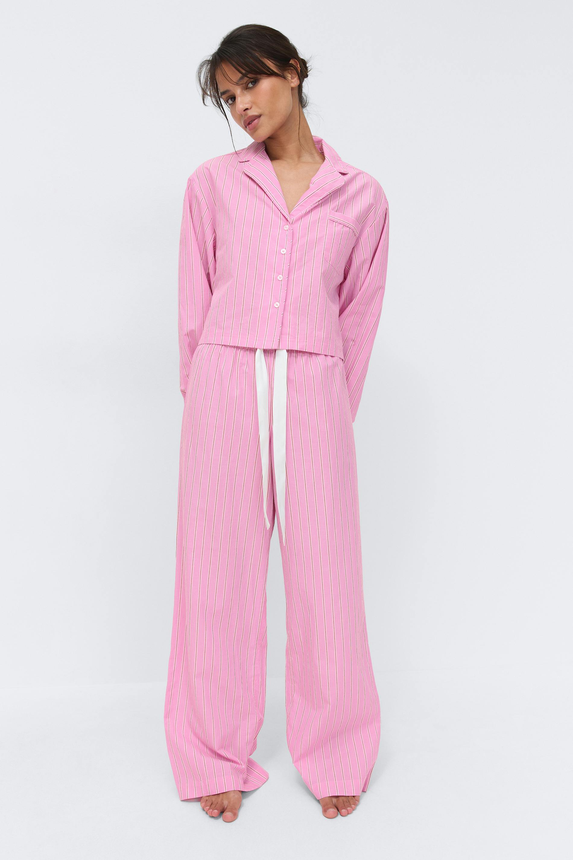 Pink vertical pinstripe two-piece pajama set: button-front long-sleeve collared shirt with chest pocket, paired with wide-leg drawstring trousers with a white ribbon tie.