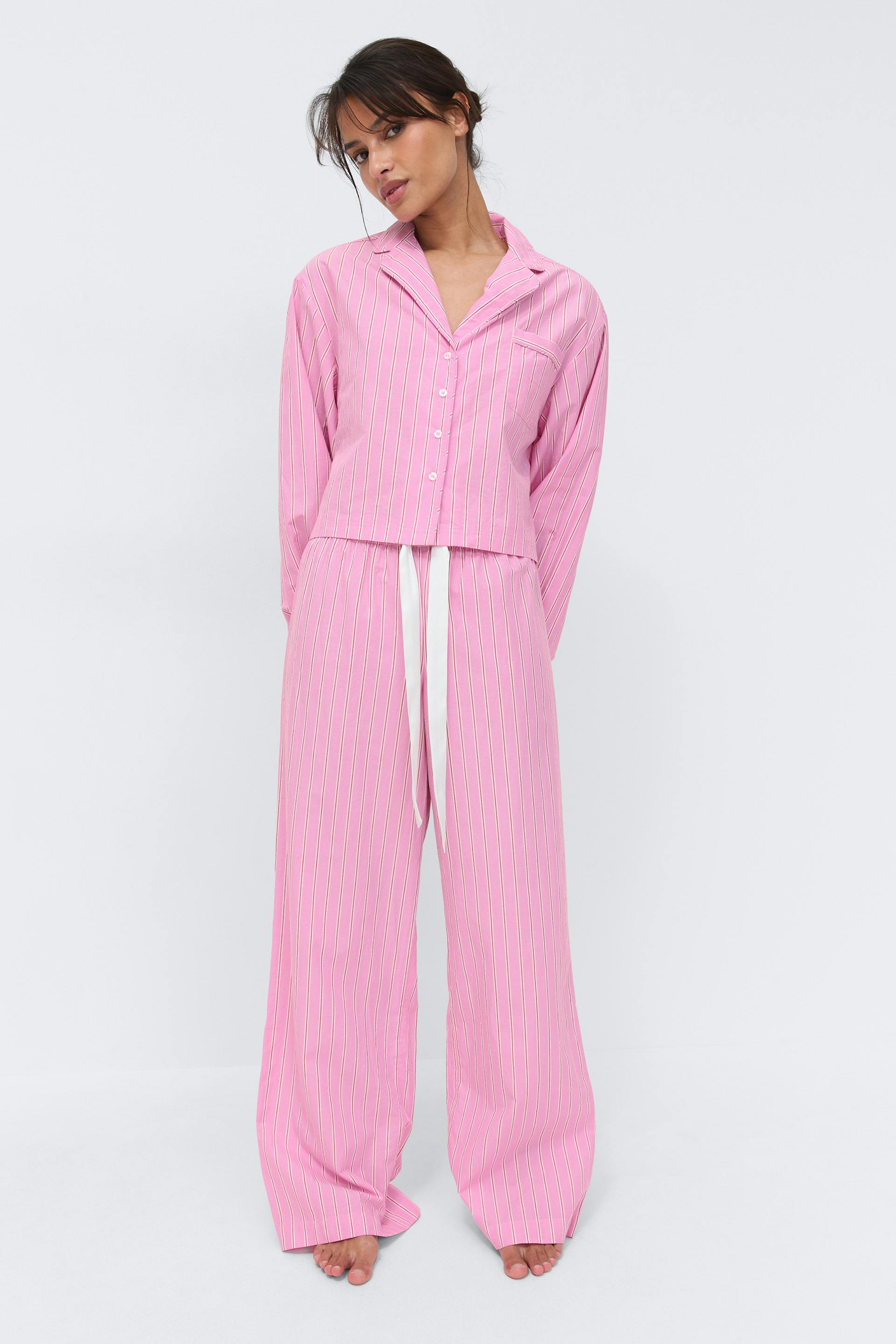 Pink vertical pinstripe two-piece pajama set: button-front long-sleeve collared shirt with chest pocket, paired with wide-leg drawstring trousers with a white ribbon tie.