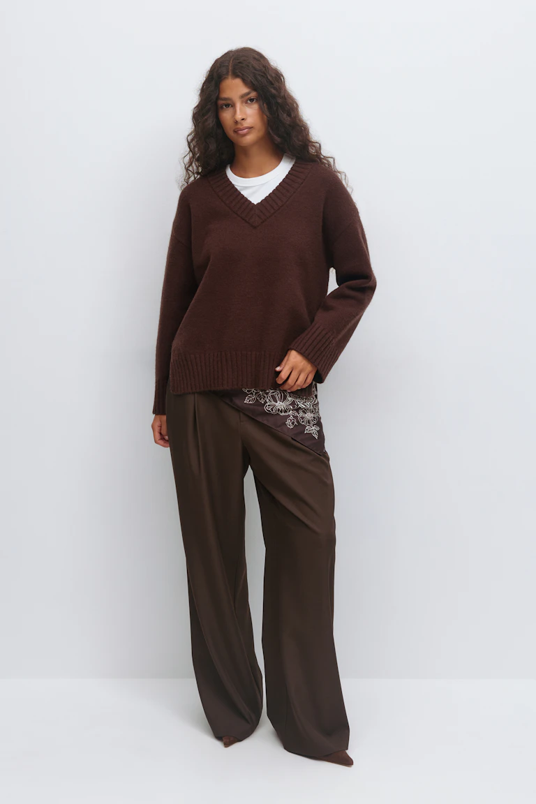 Weißes T-Shirt mit Rundhalsausschnitt unter einem dunkelbraunen gerippten Pullover mit V-Ausschnitt, gepaart mit einer dunkelbraunen Hose mit weitem Bein, die an der Hüfte einen braunen, mit Blumen bestickten Seideneinsatz aufweist, und braunen, spitz zulaufenden Schuhen.