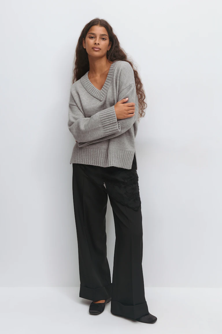 Grå oversized sweater med V-udskæring og ribkant sammen med sorte bukser med brede ben og manchetter, en sort underkjole med blondekant indenunder og sorte flade sko med firkantede tæer.