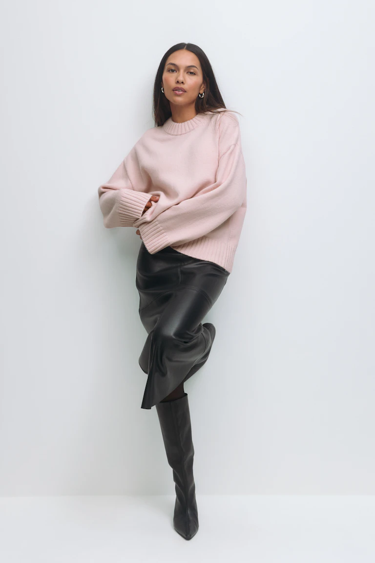 Femme portant un pull rose pâle surdimensionné à col ras du cou, une jupe midi en cuir noir et des bottes noires à bout pointu jusqu'au genou.