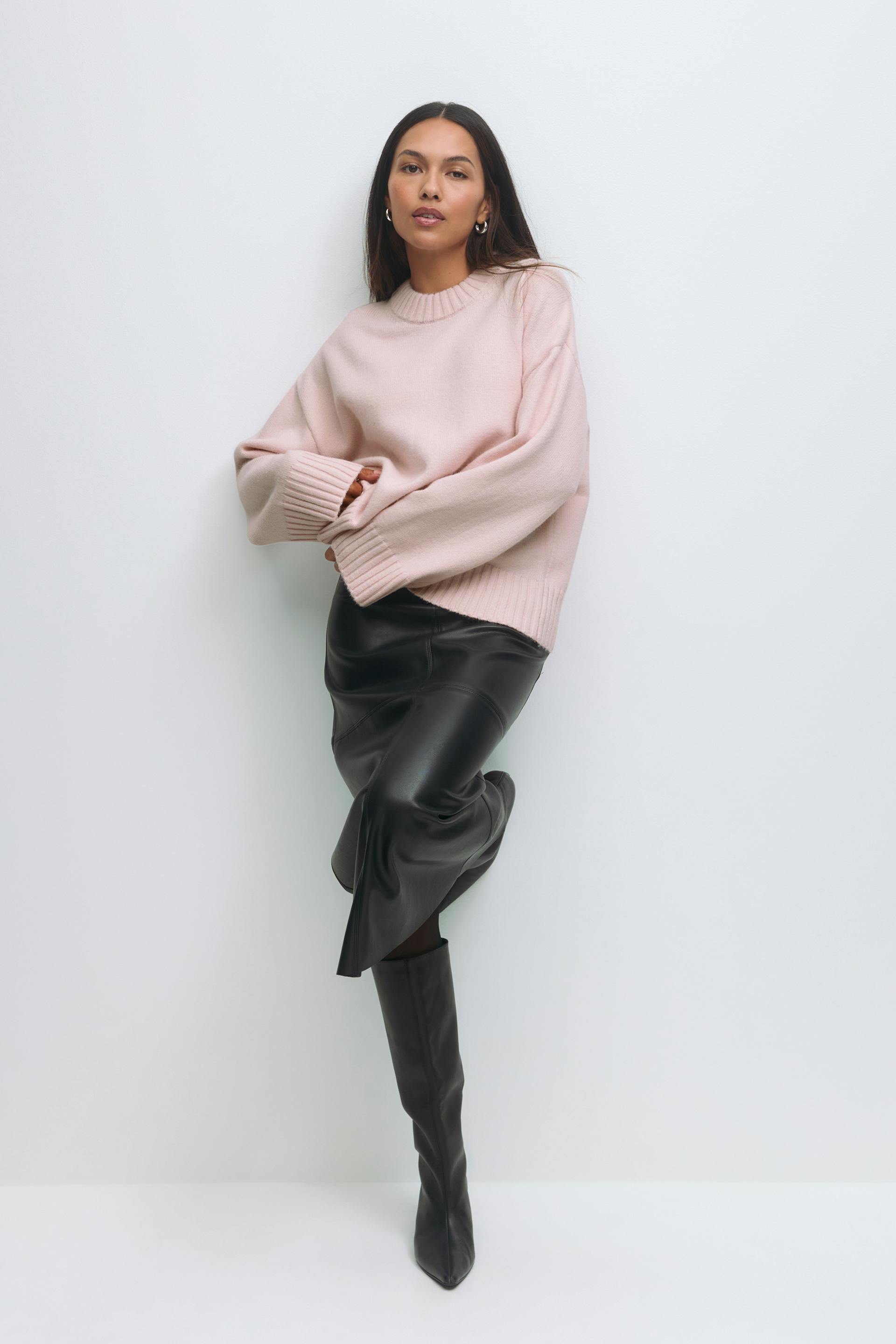 Vrouw draagt een lichtroze oversized geribde trui met ronde hals, zwarte leren midirok en zwarte kniehoge puntschoenen.