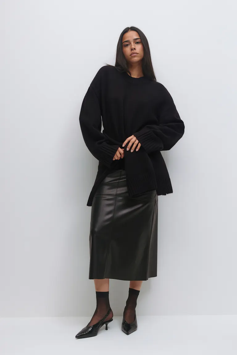 Schwarzer Pullover mit übergroßem Rundhalsausschnitt und gerippten Bündchen und Saum, schwarzer Midirock aus Leder, schwarze durchsichtige Söckchen und spitze schwarze Slingback-Kitten-Heels.