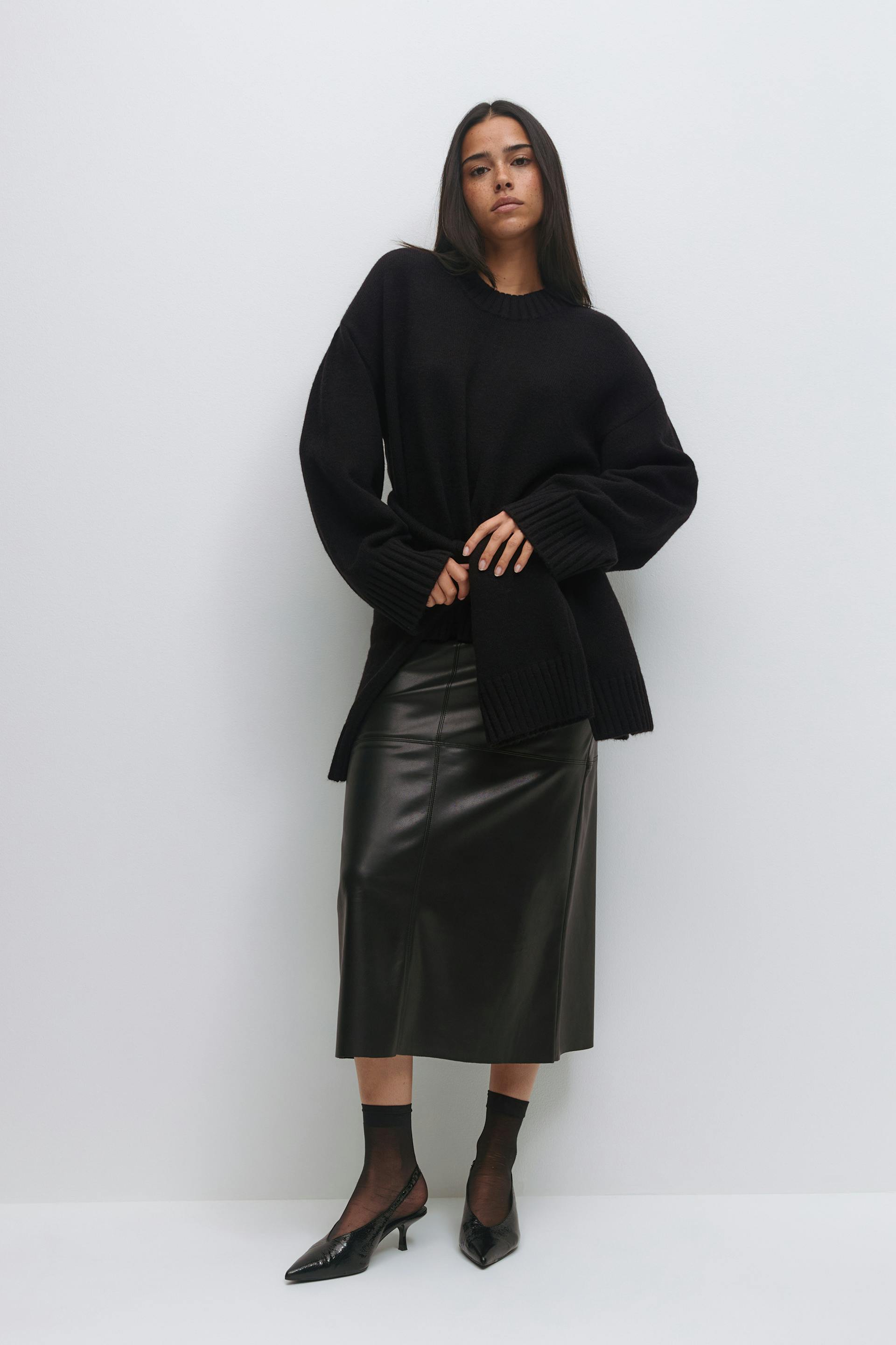 Oversized zwarte crewneck-trui met geribde boorden en zoom, zwarte leren midi-rok, transparante zwarte enkelsokken en spitse zwarte slingback-kittenhakken.
