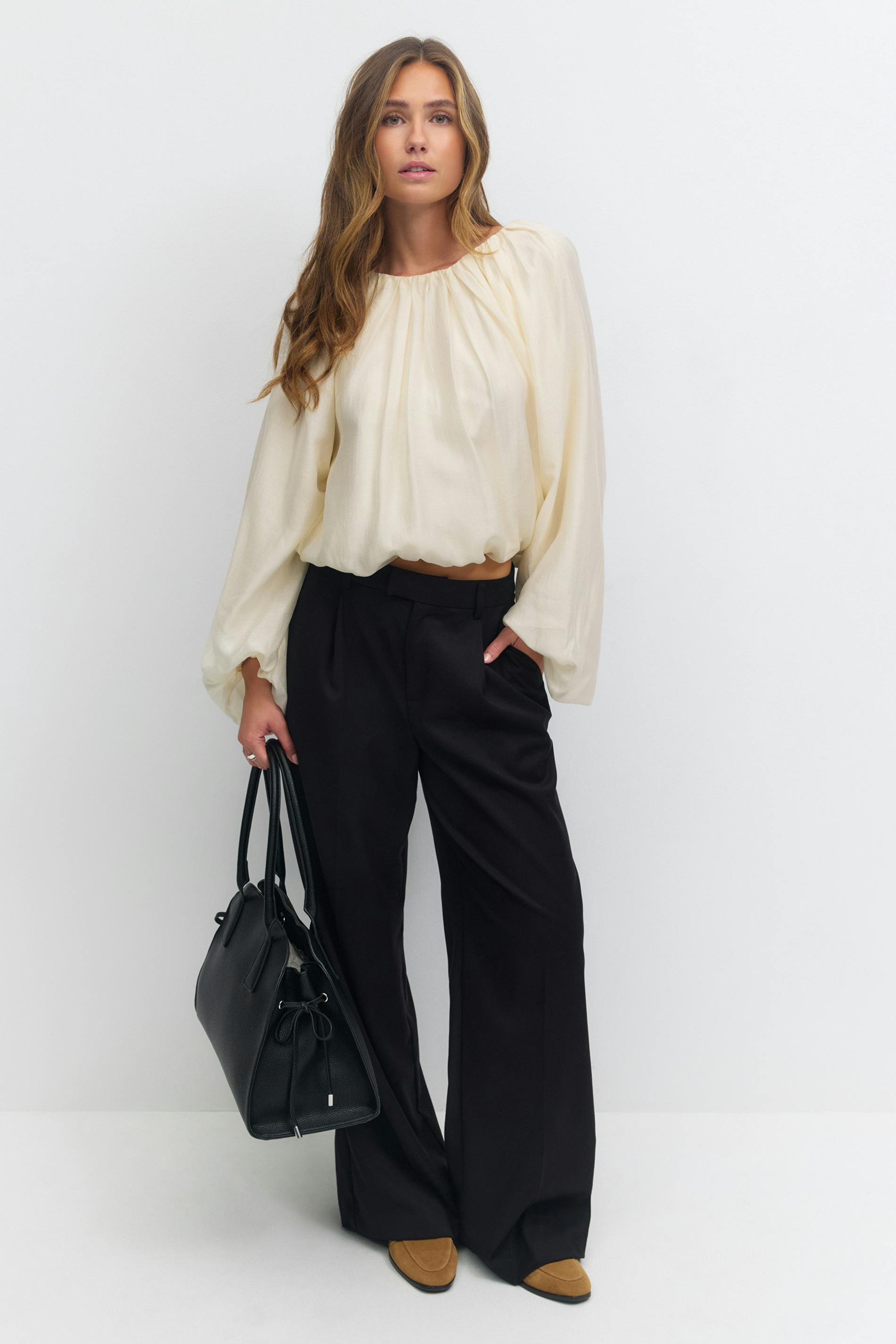 Kremfarget, langermet bluse med rynkete utringning, bølgende ermer og blousonkant; svarte bukser med høy midje og vide ben; brunfargede loafers i semsket skinn; svart strukturert skinnveske.