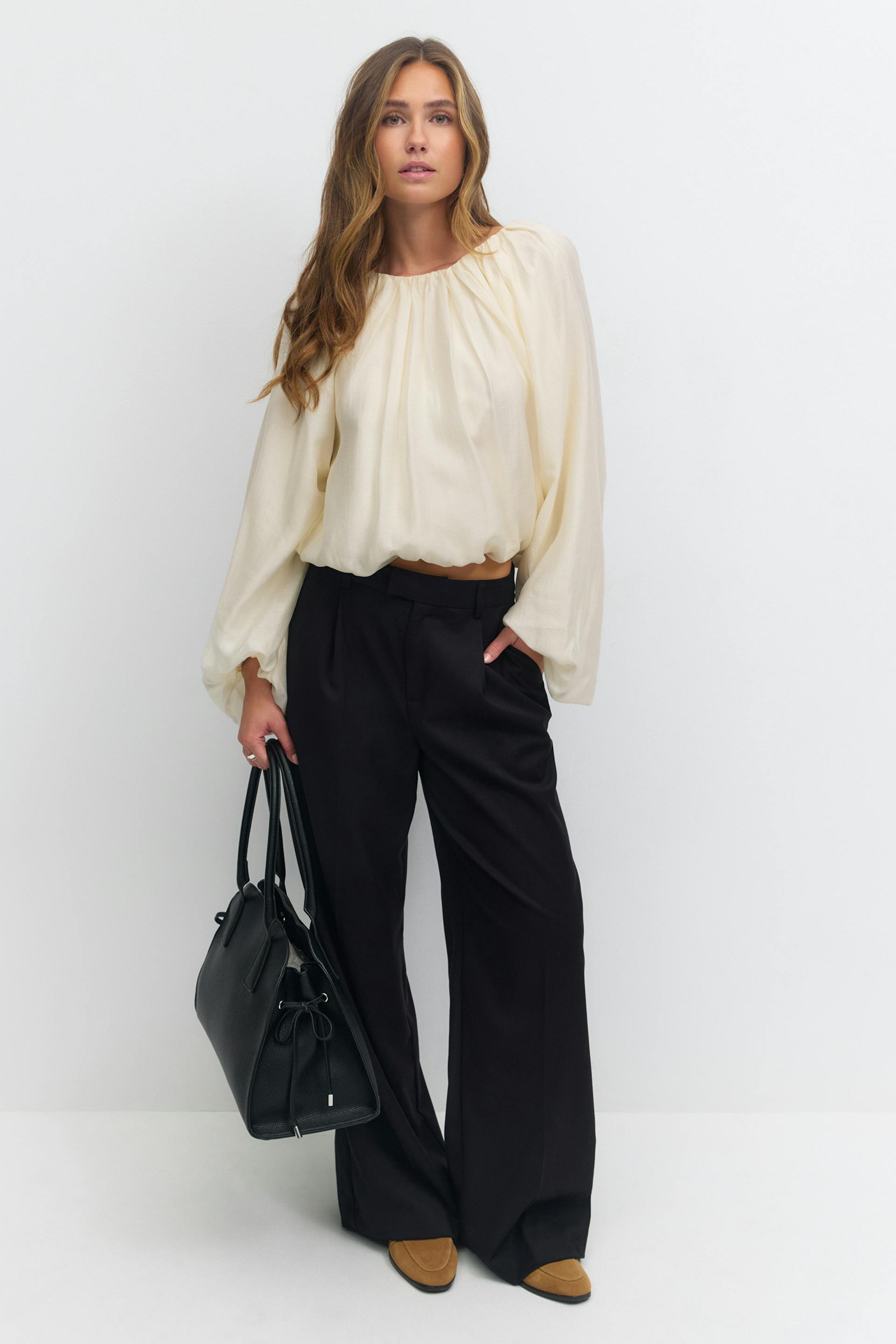 Kremfarget, langermet bluse med rynkete utringning, bølgende ermer og blousonkant; svarte bukser med høy midje og vide ben; brunfargede loafers i semsket skinn; svart strukturert skinnveske.