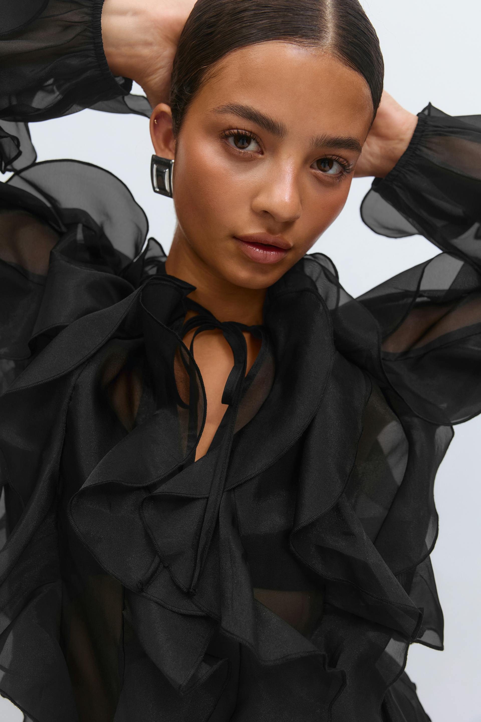 Black sheer ruffled chiffon blouse with tie neckline, paired with black satin drawstring trousers and silver rectangular statement earrings.
