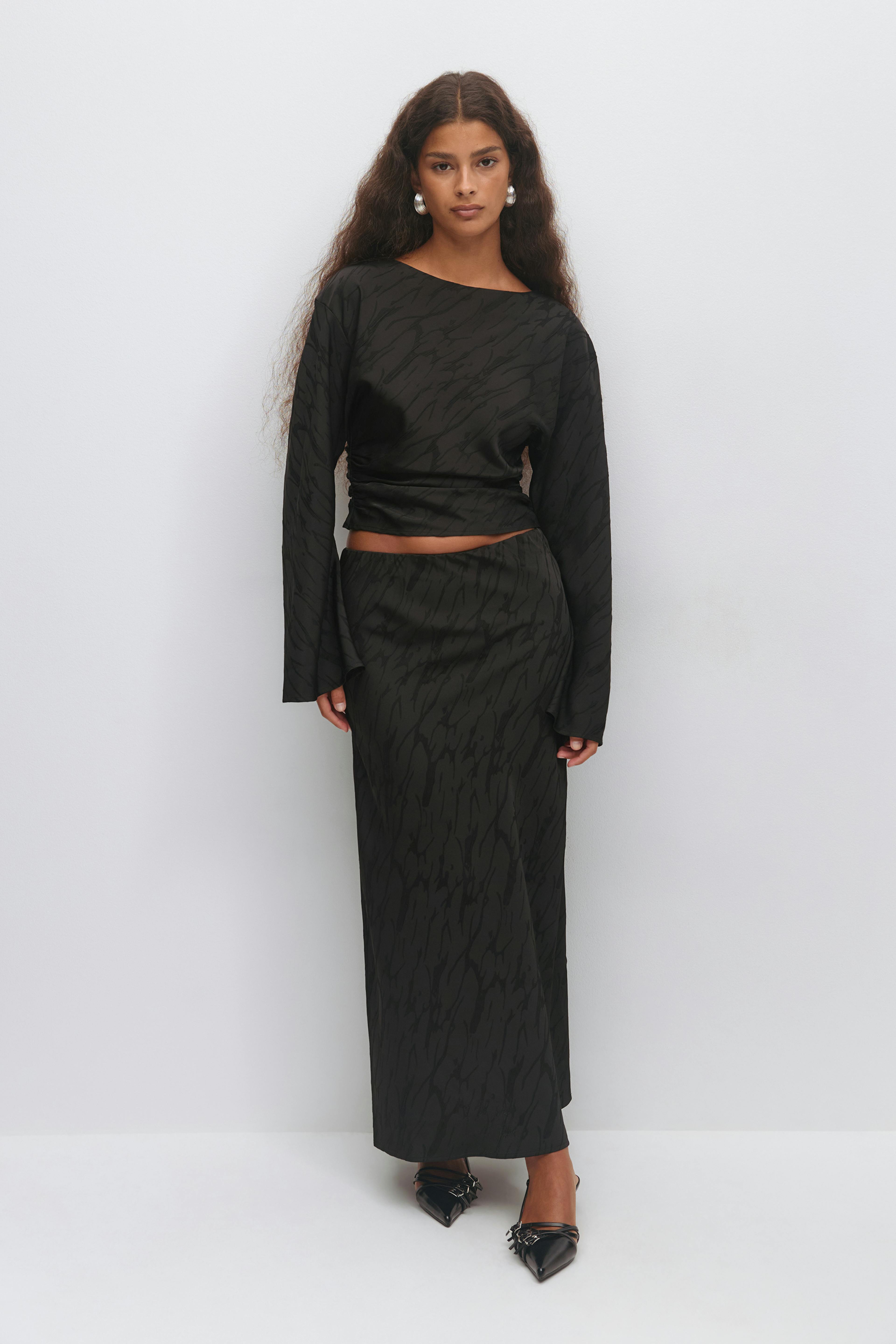 Black two-piece set: long-sleeve cropped top with subtle textured abstract print and matching high-waist, ankle-length straight skirt; paired with black pointed-toe strappy heels with metal detailing and silver statement hoop earrings.