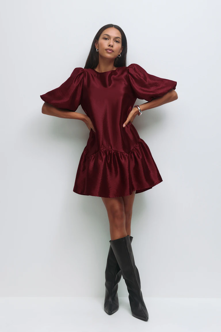 Burgundy satin mini dress with voluminous puff sleeves and a gathered ruffle hem; black knee‑high pointed leather boots; silver hoop earrings and a silver bracelet.